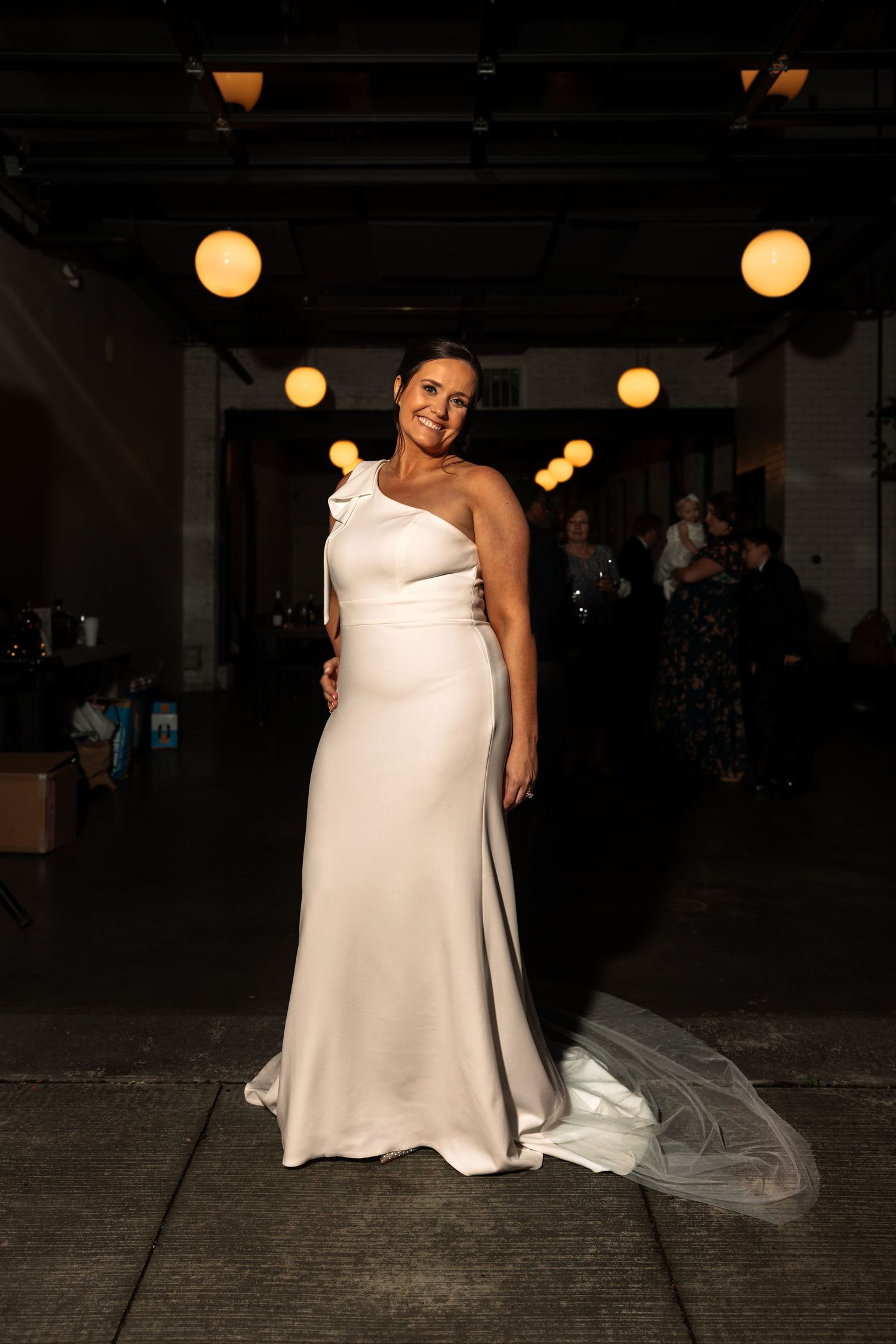 Elegant bride in a one-shoulder white gown with cathedral veil at reception.