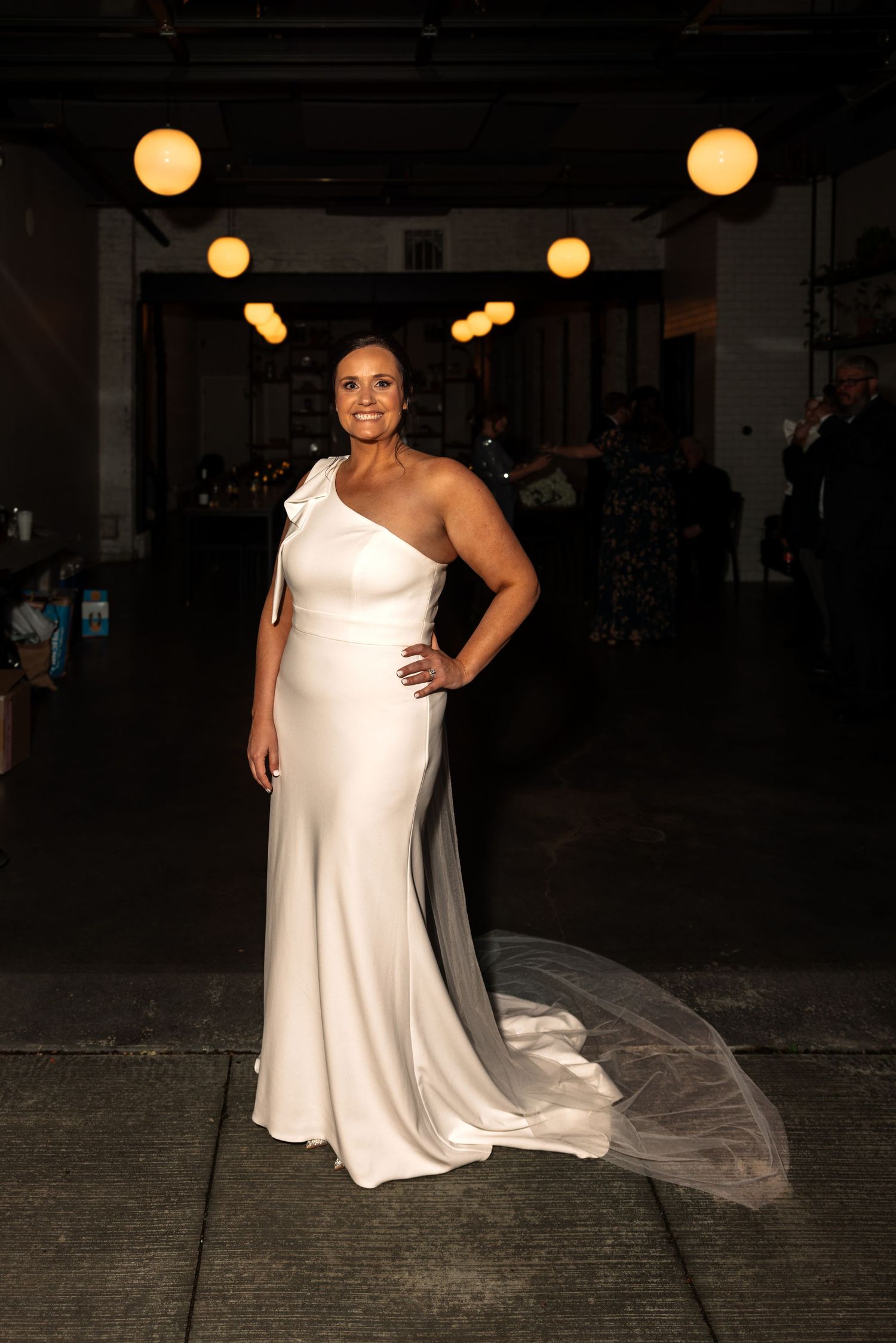 Smiling bride in a fitted one-shoulder white satin gown with cathedral veil.