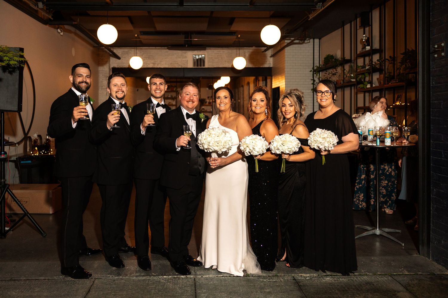 Wedding party group photo with groomsmen in tuxedos and bridesmaids in black gowns holding white bouquets.