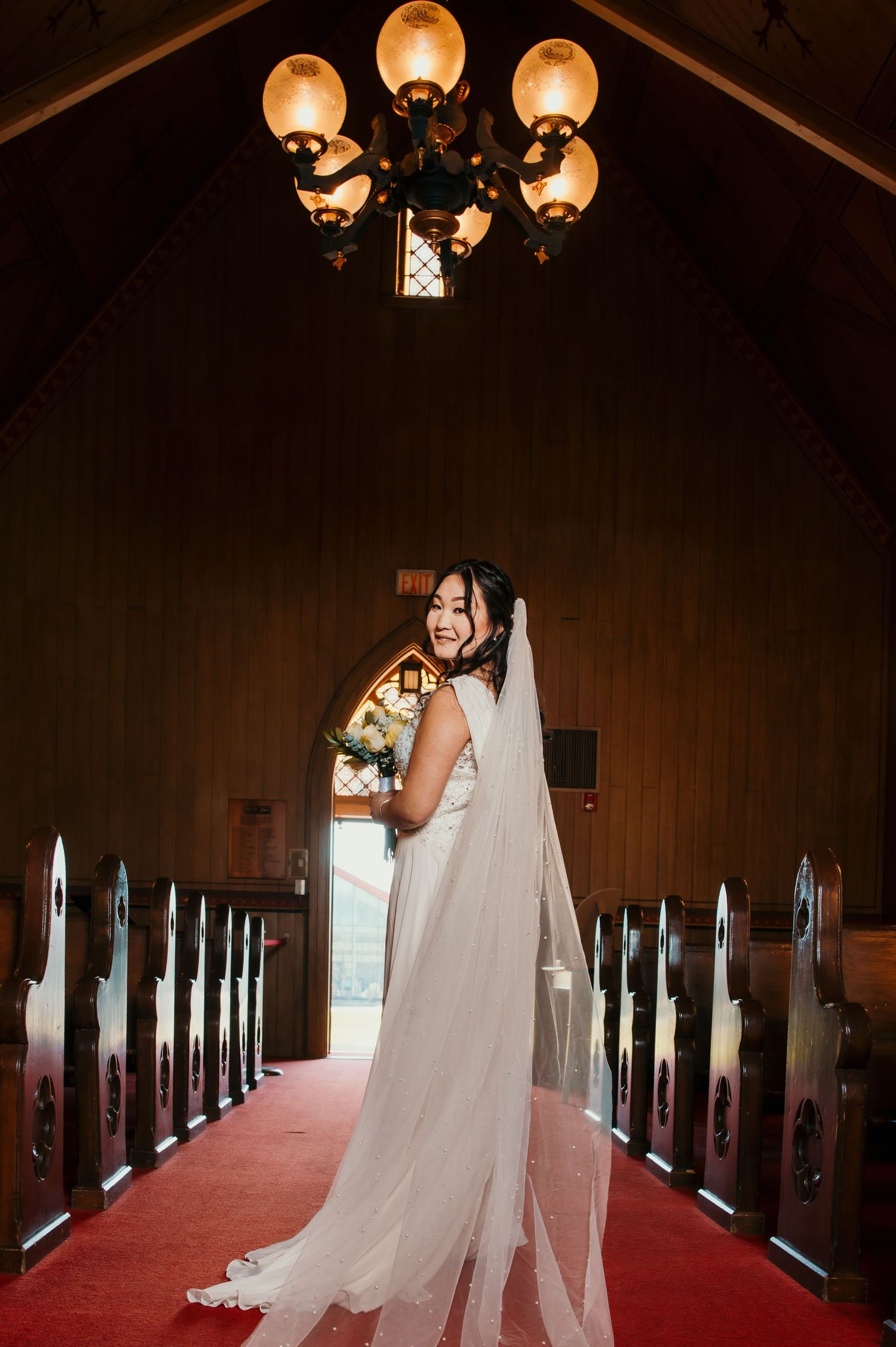 Joyful bride in white gown with long veil laughing while holding bouquet in chapel aisle.
