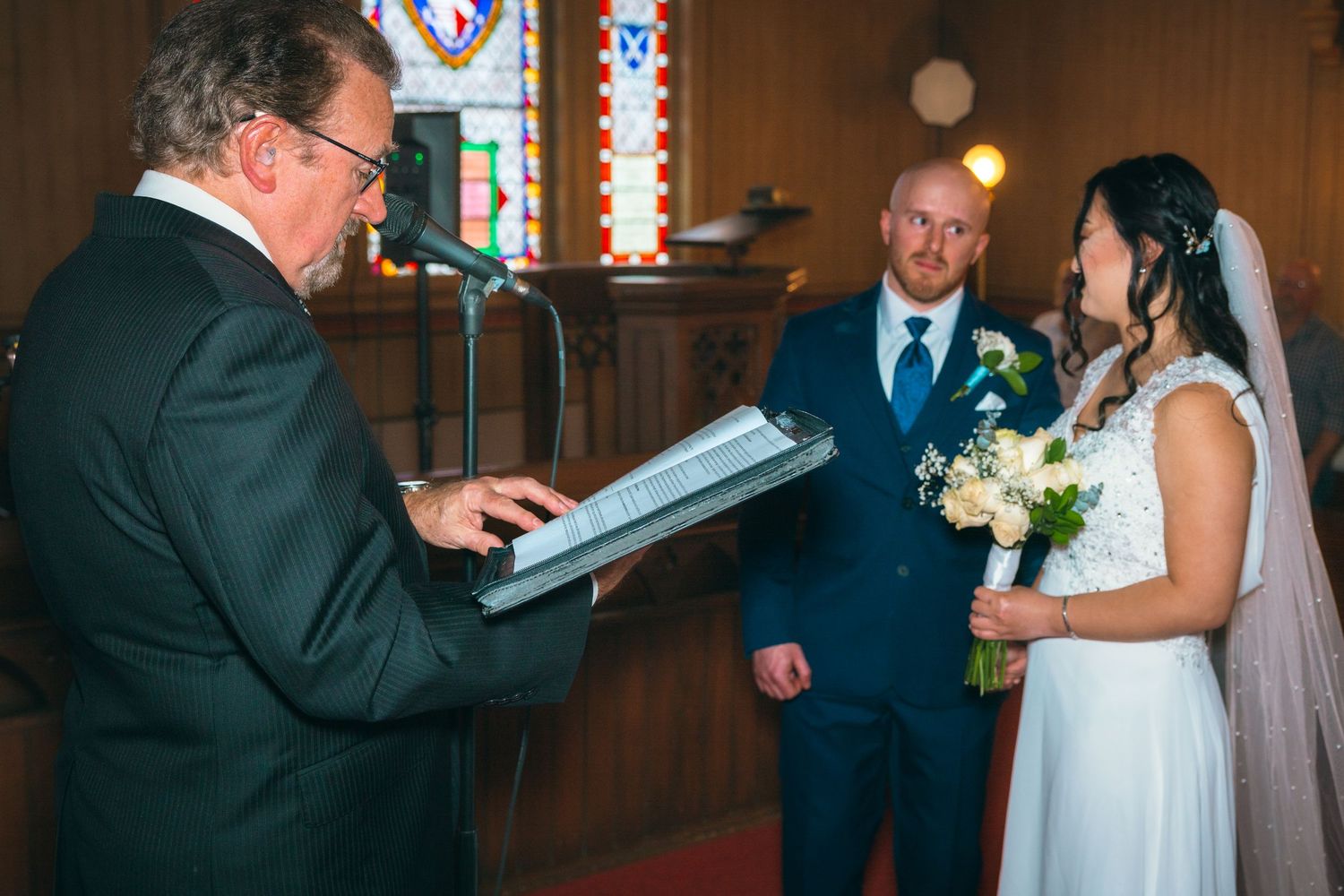 Wedding officiant reads ceremony script as bride with bouquet and groom stand at church altar.