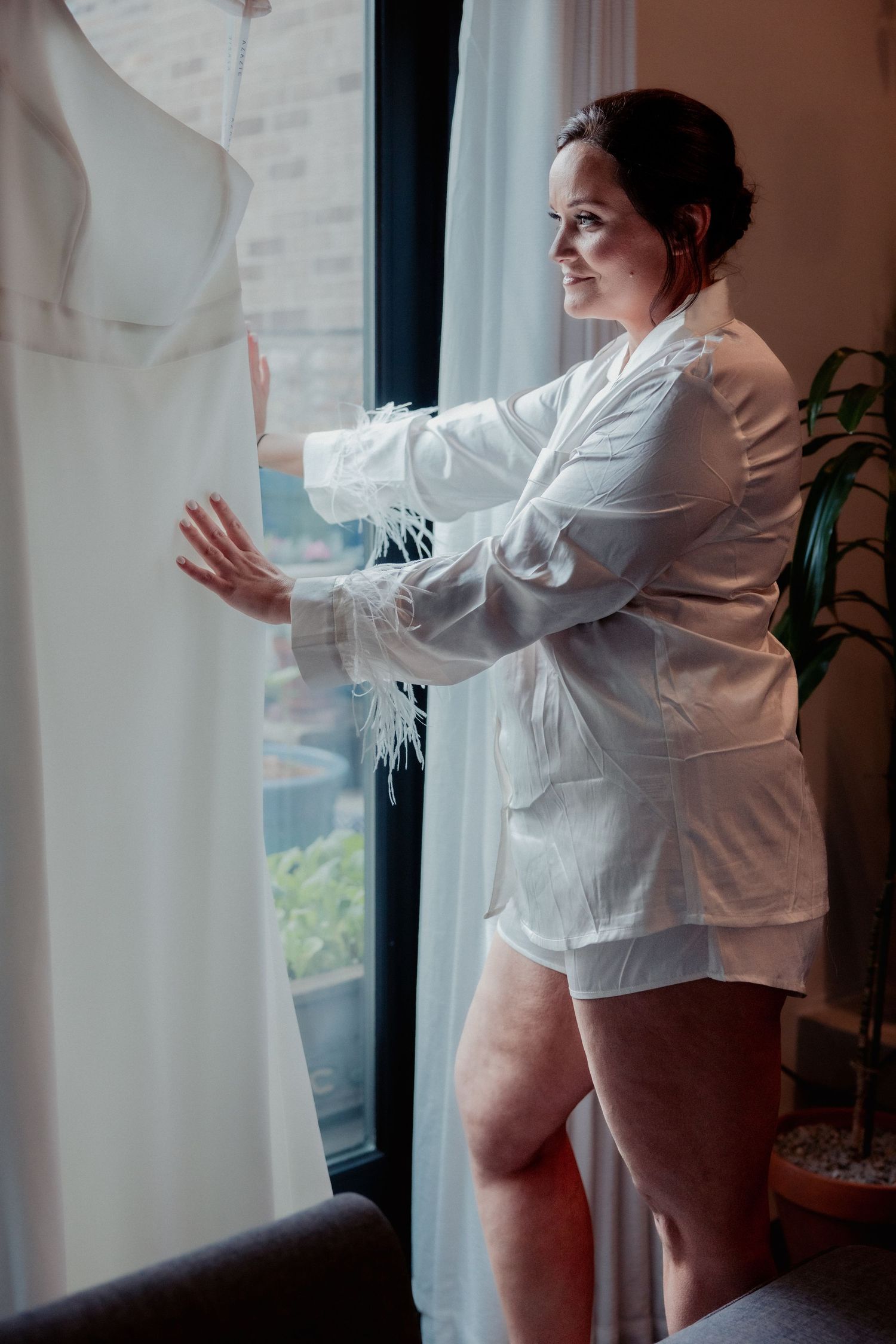 Bride in white feather robe standing by window touching her hanging wedding gown.