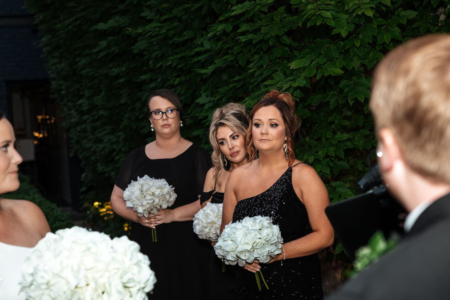 Three bridesmaids in black dresses holding white hydrangea bouquets watching outdoor wedding ceremony.