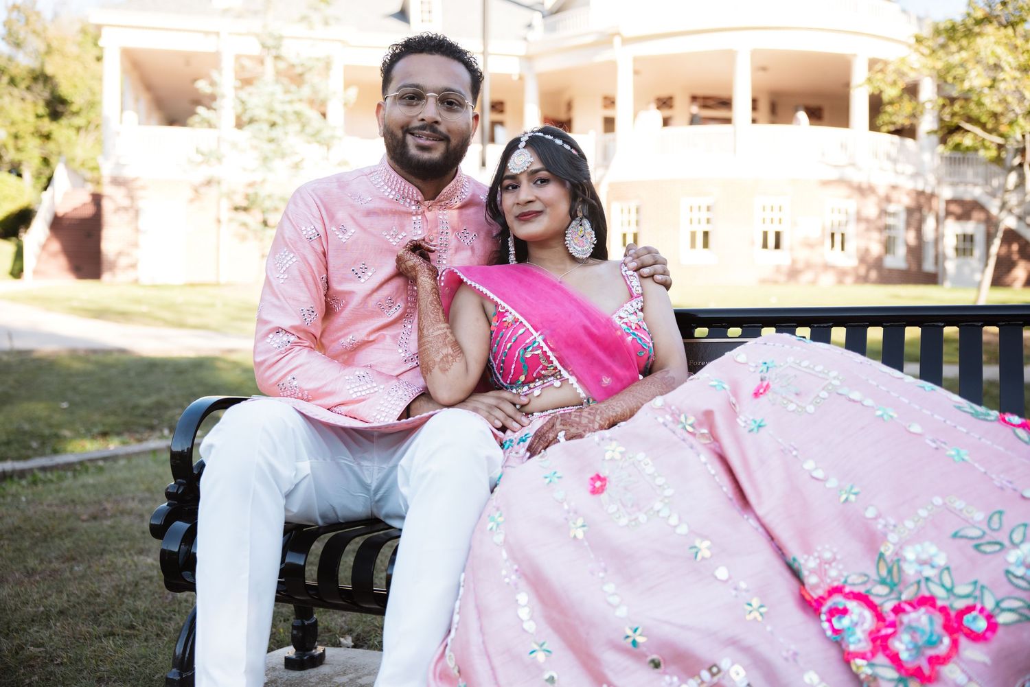 Indian couple in coordinating pink outfits sitting together on outdoor bench smiling at a grand estate.