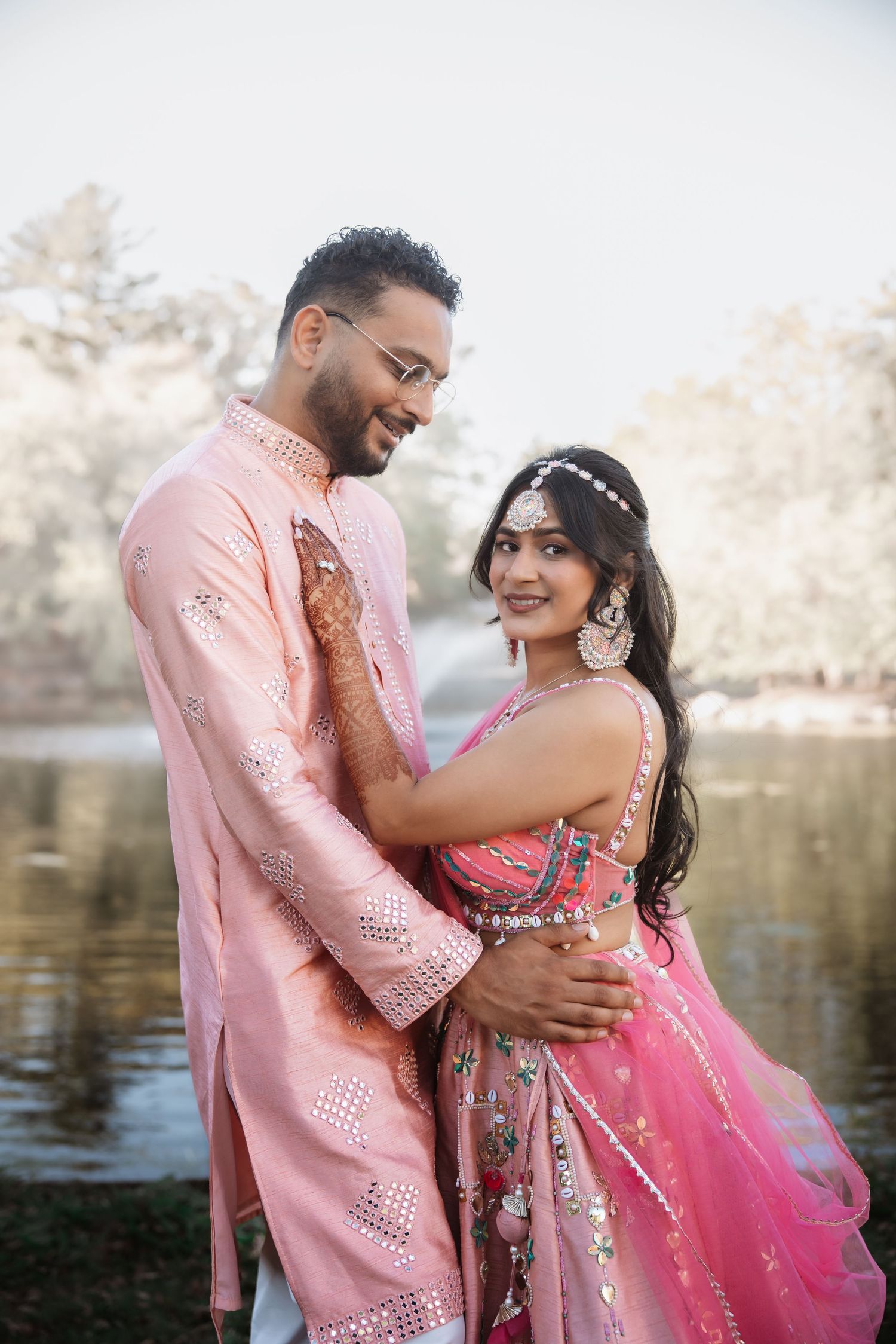 Indian couple in pink traditional attire embracing romantically near a serene lake.