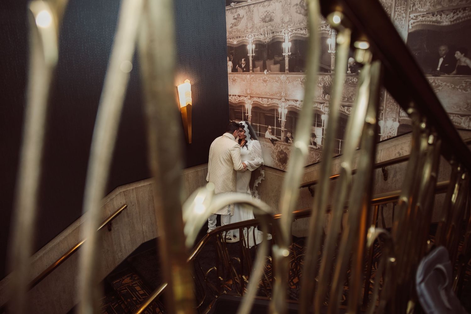 Artistic shot of wedding couple embracing on staircase framed by golden candelabras in dimly lit venue.