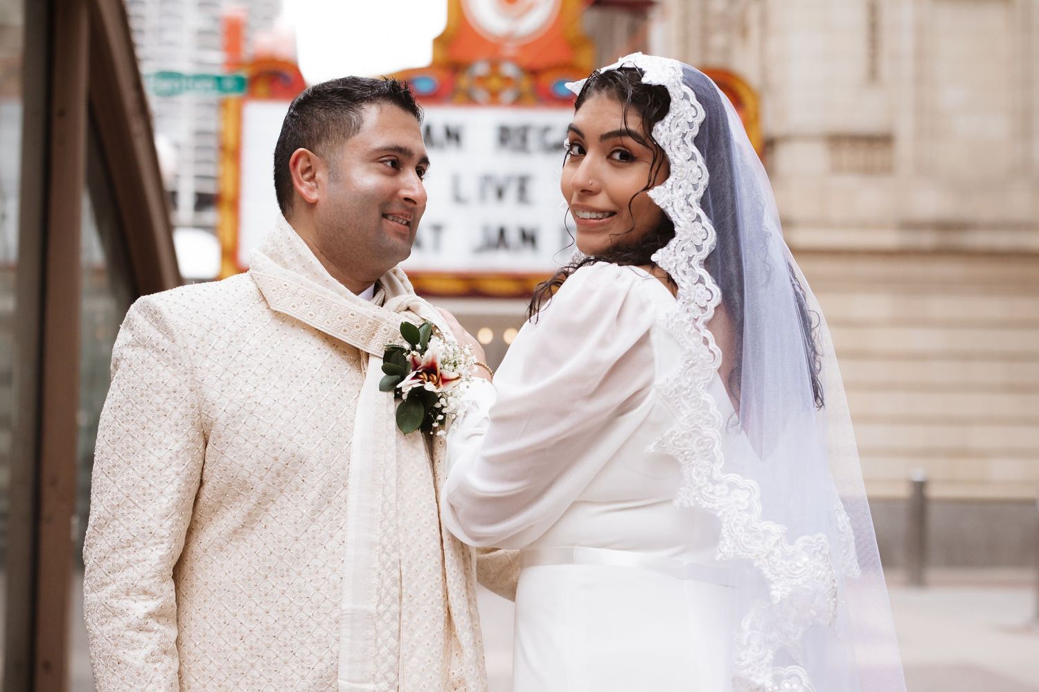 Indian newlyweds in white wedding attire share a romantic glance outside Chicago Theatre.