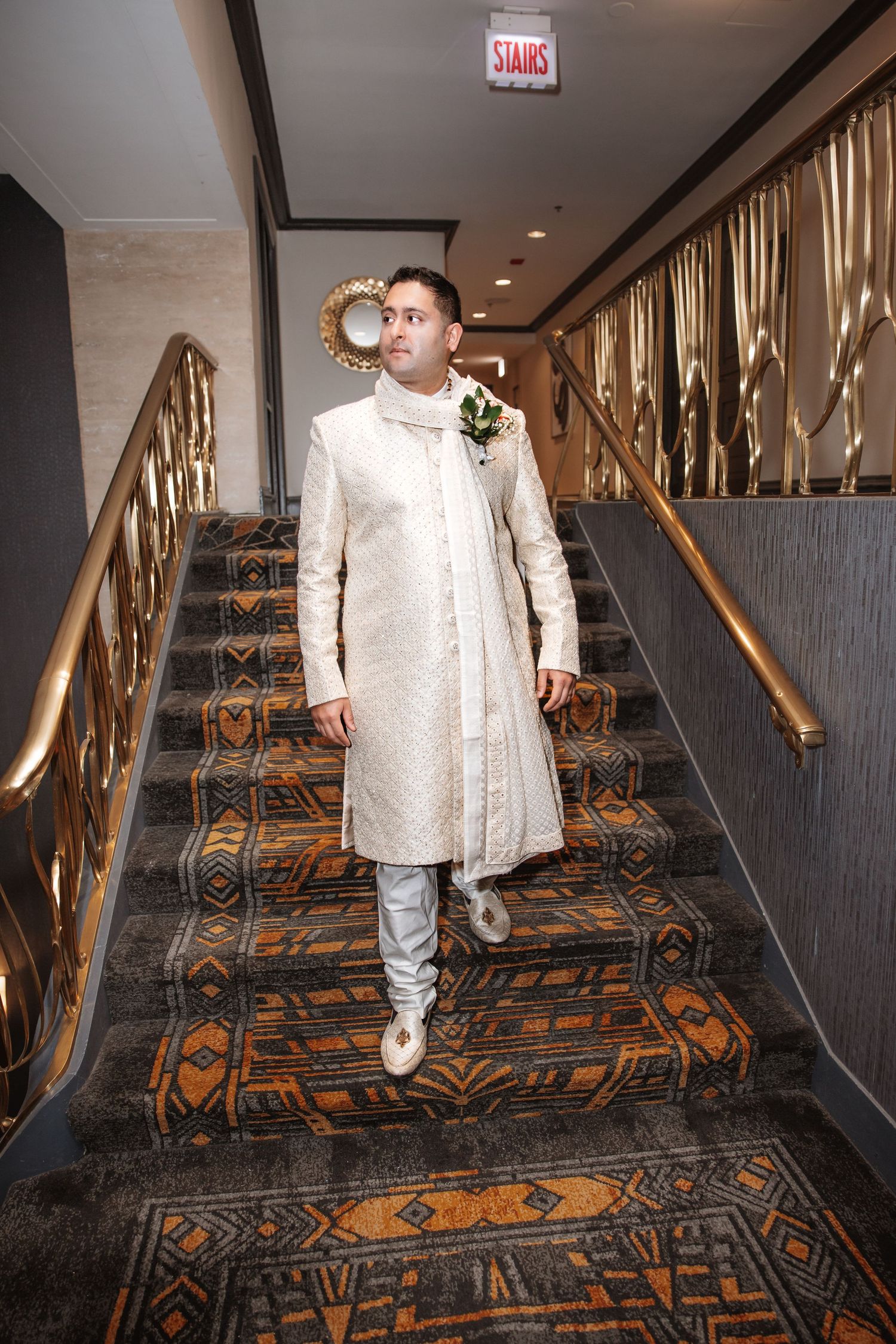 Groom in traditional ivory Indian sherwani with boutonniere descending ornate carpeted staircase in elegant hotel venue.
