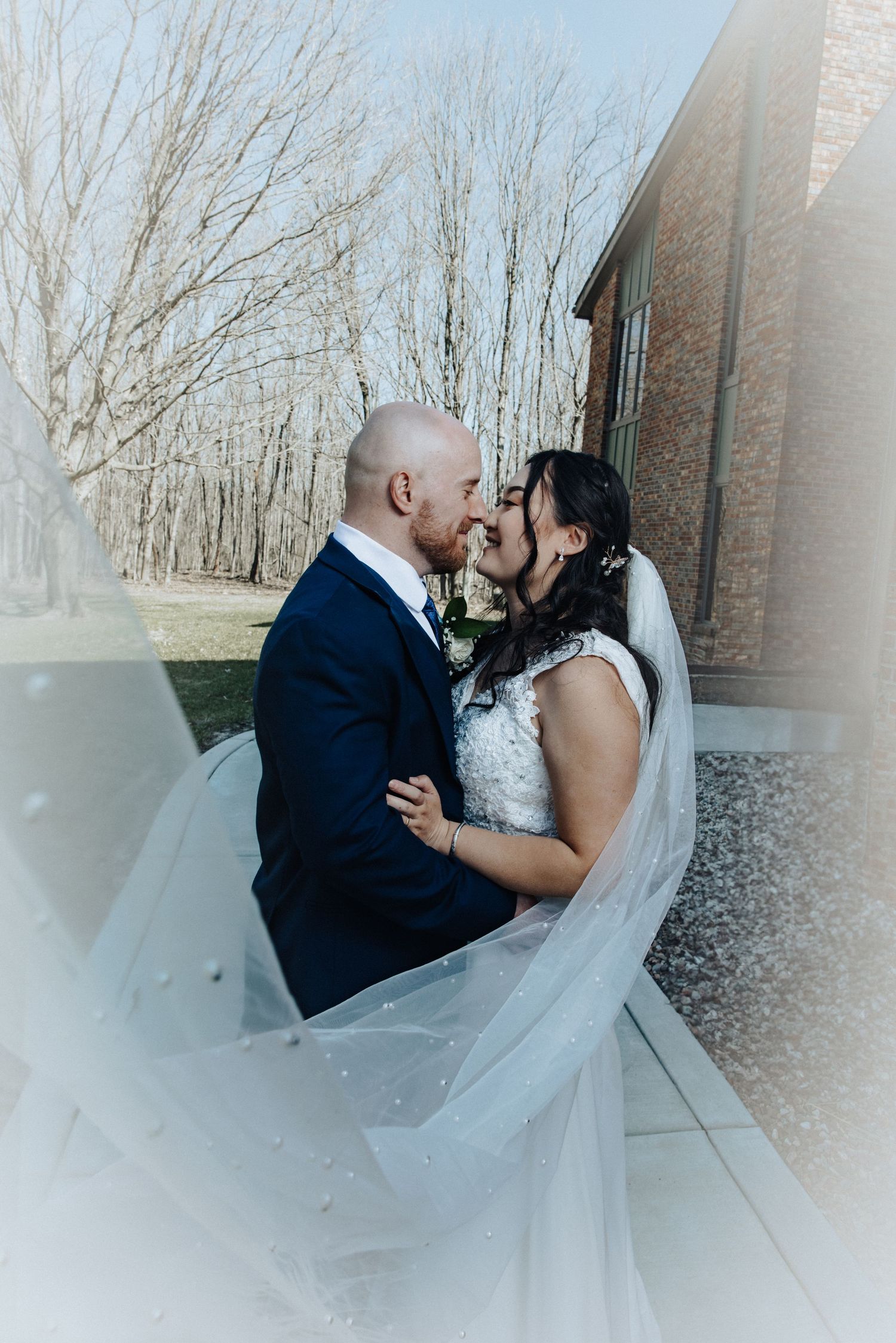 Newlyweds share a tender moment nose-to-nose with wedding veil billowing around them near a brick venue.