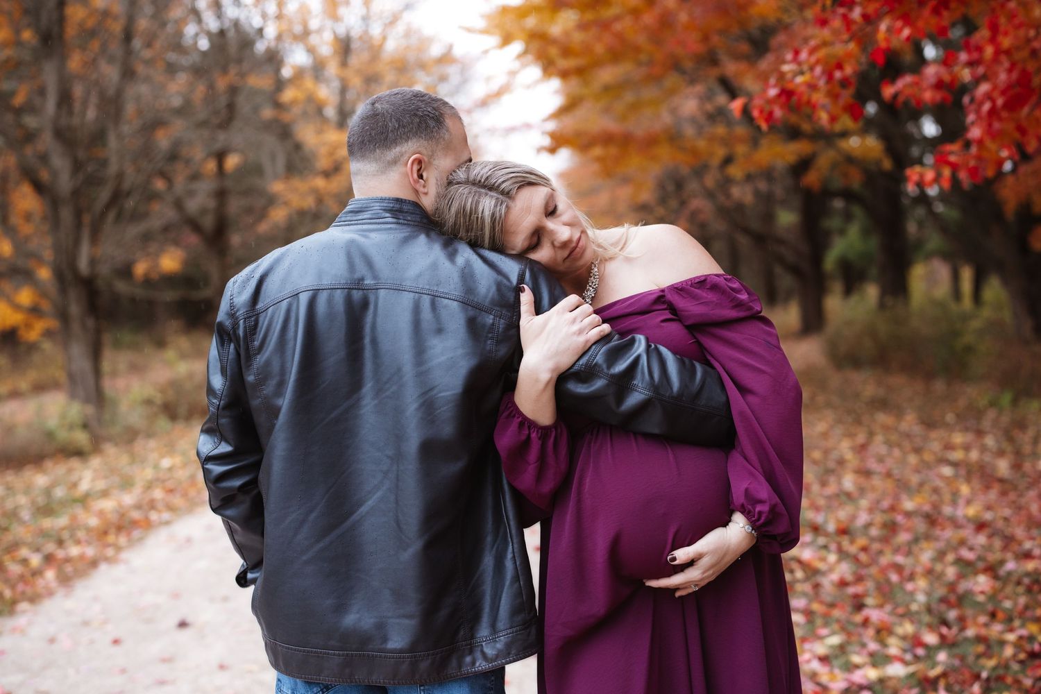 Couple sharing tender moment on autumn path, pregnant woman in purple gown leans on mans shoulder in leather jacket.