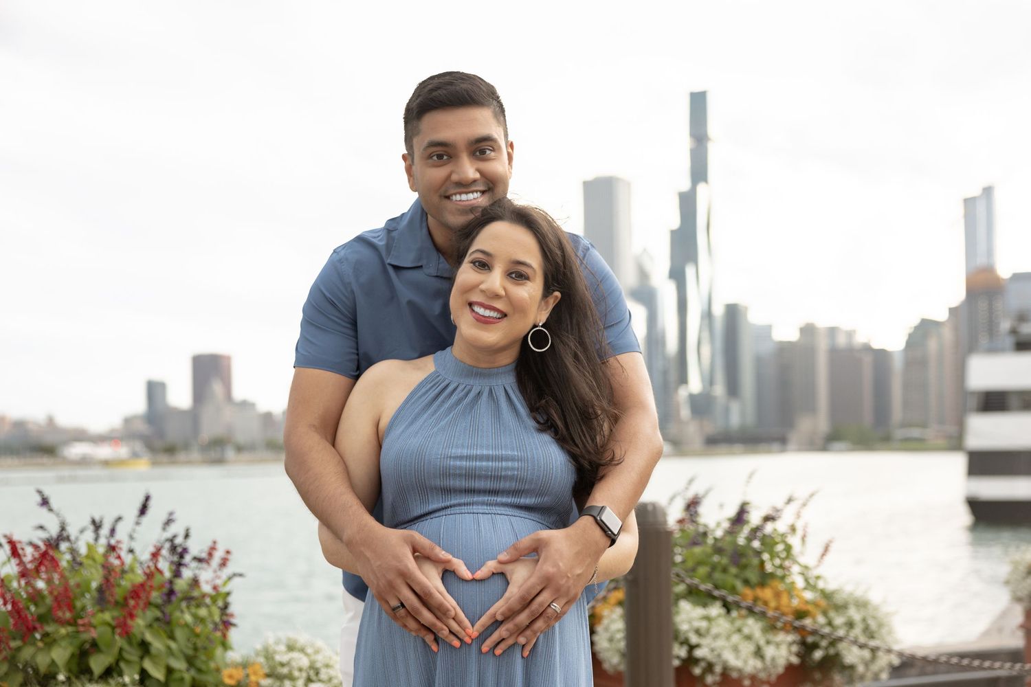 Pregnant couple posing with heart hands on baby bump with Chicago skyline and waterfront in background.