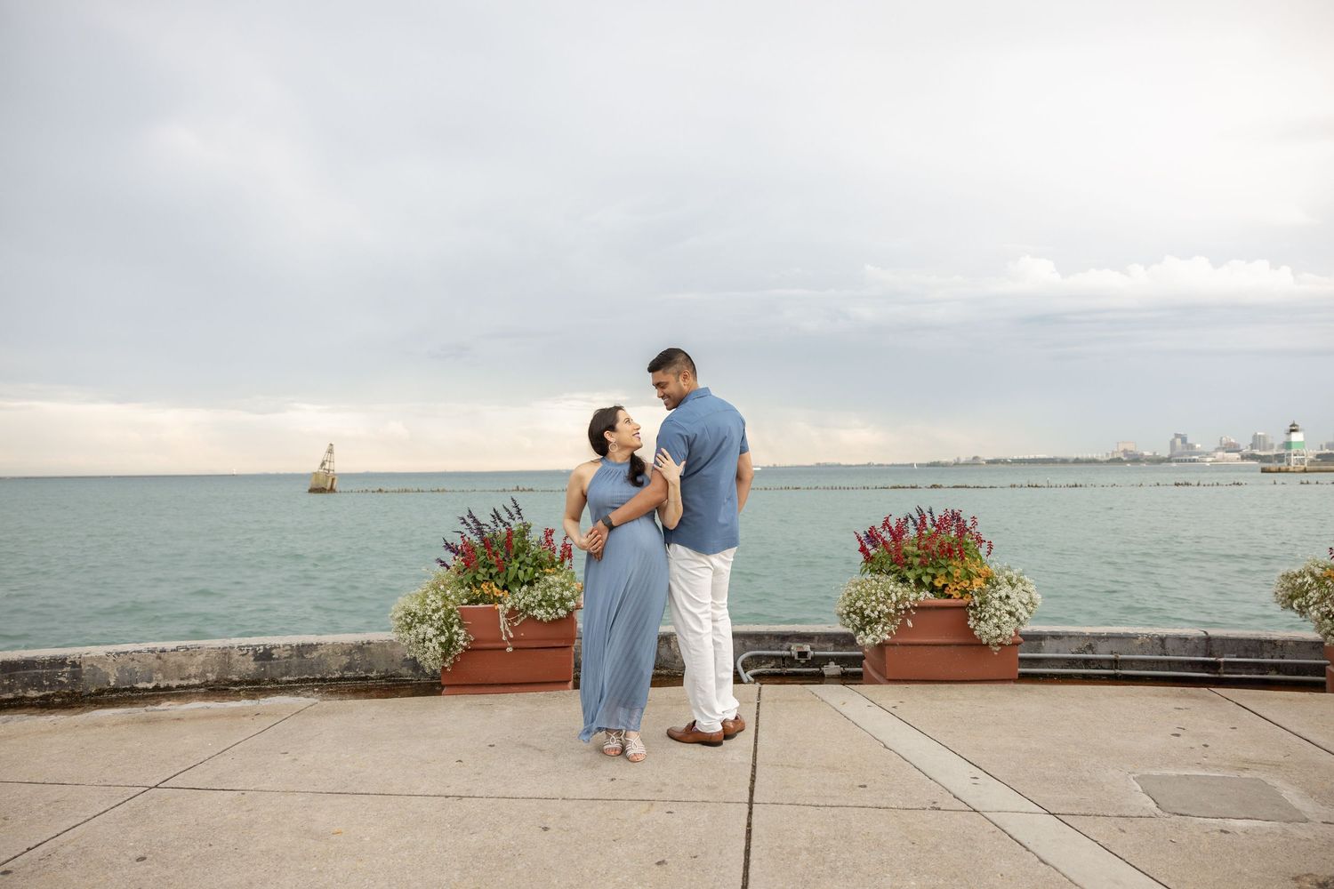 Couple standing together on waterfront terrace with sailboat in background, woman in blue maternity dress, cloudy sky.