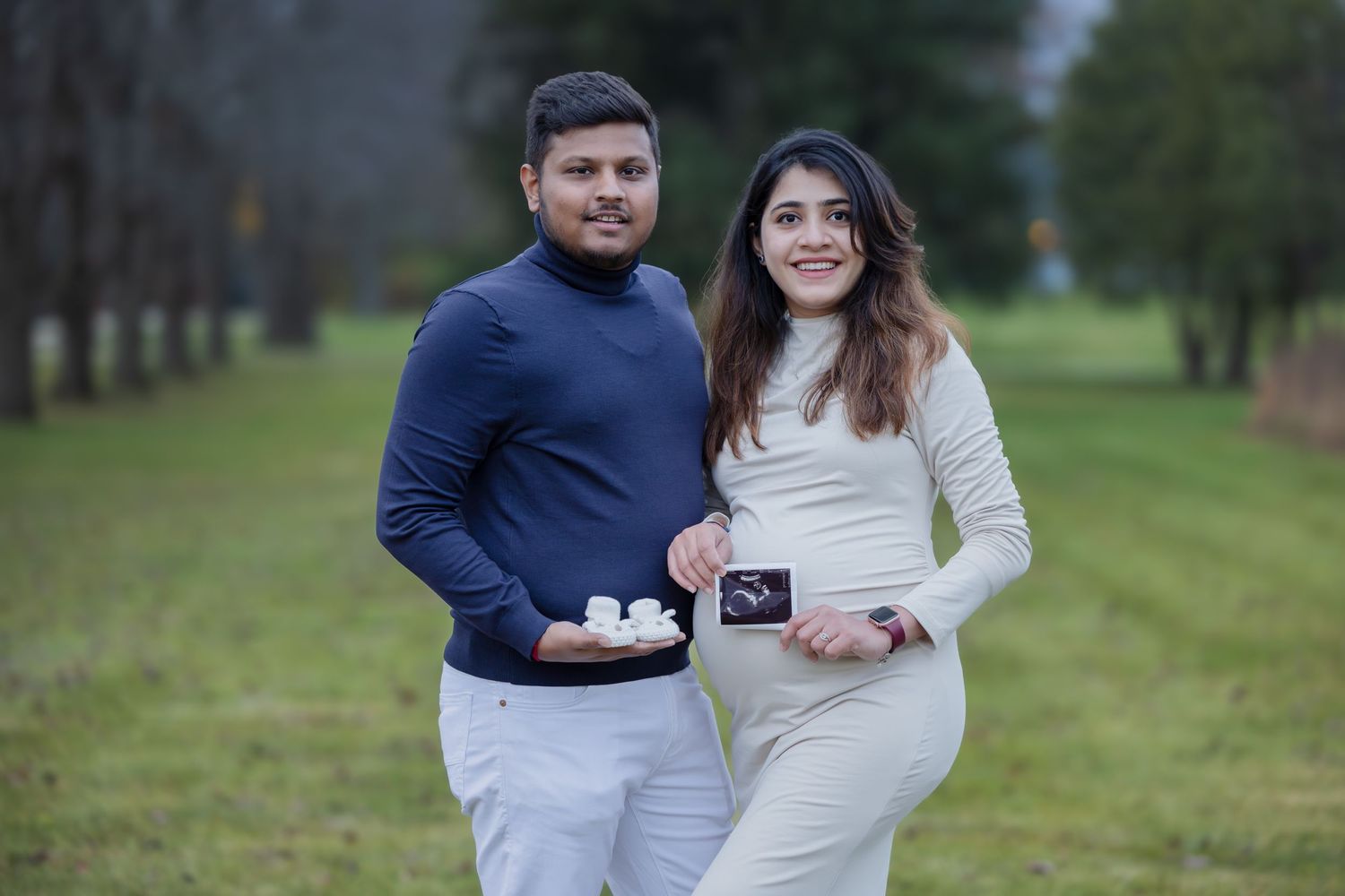 Expecting couple displaying baby shoes and ultrasound images in a park setting.