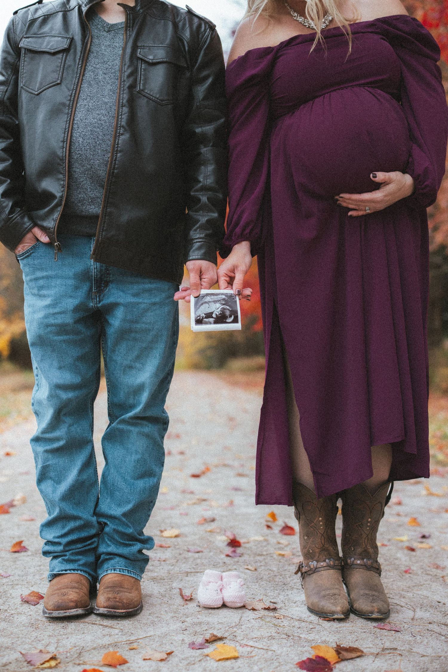 Couple holding ultrasound photo during fall maternity shoot with tiny pink baby shoes on ground.