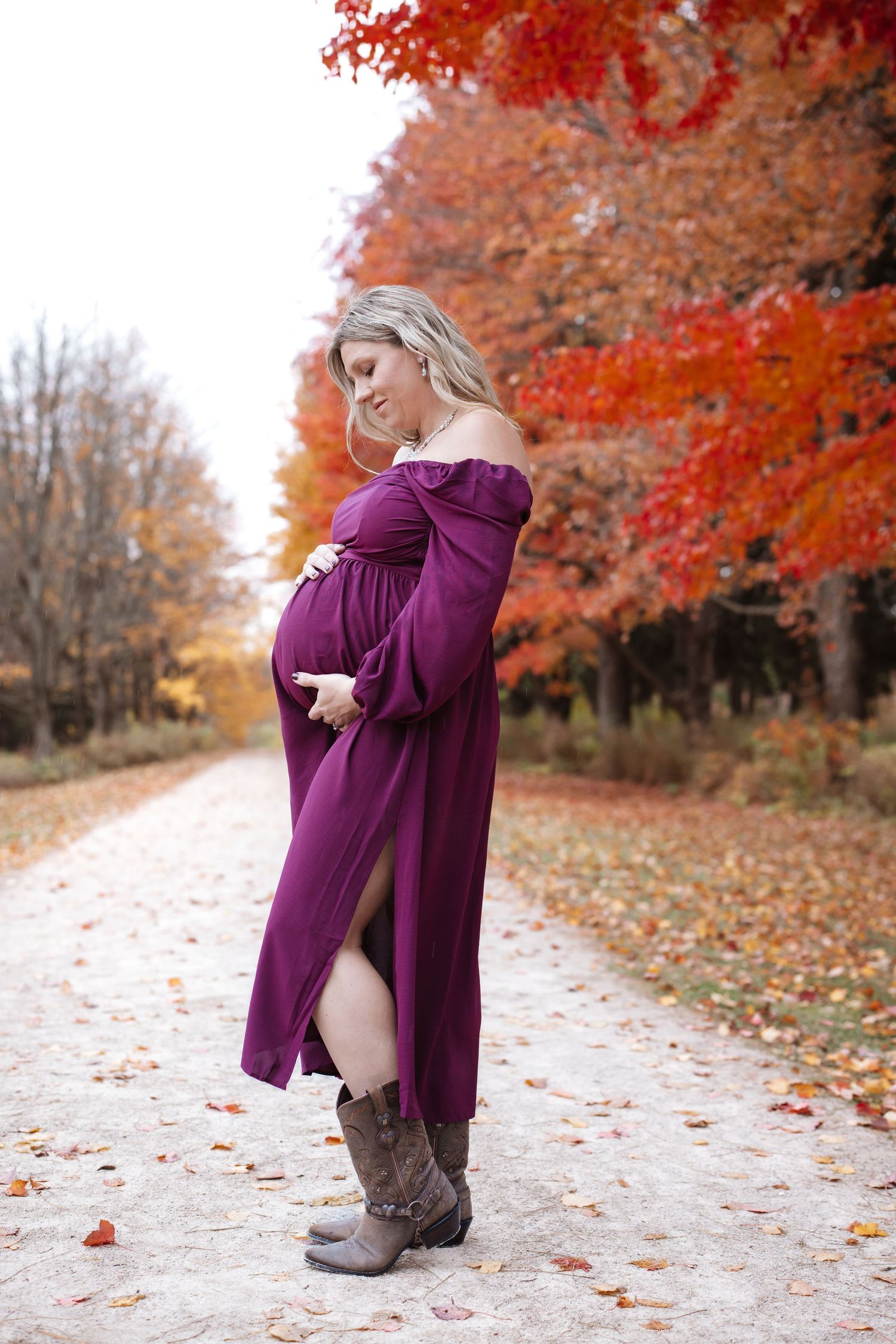 Pregnant woman in purple off-shoulder dress and cowboy boots posing on fall leaf-covered path.
