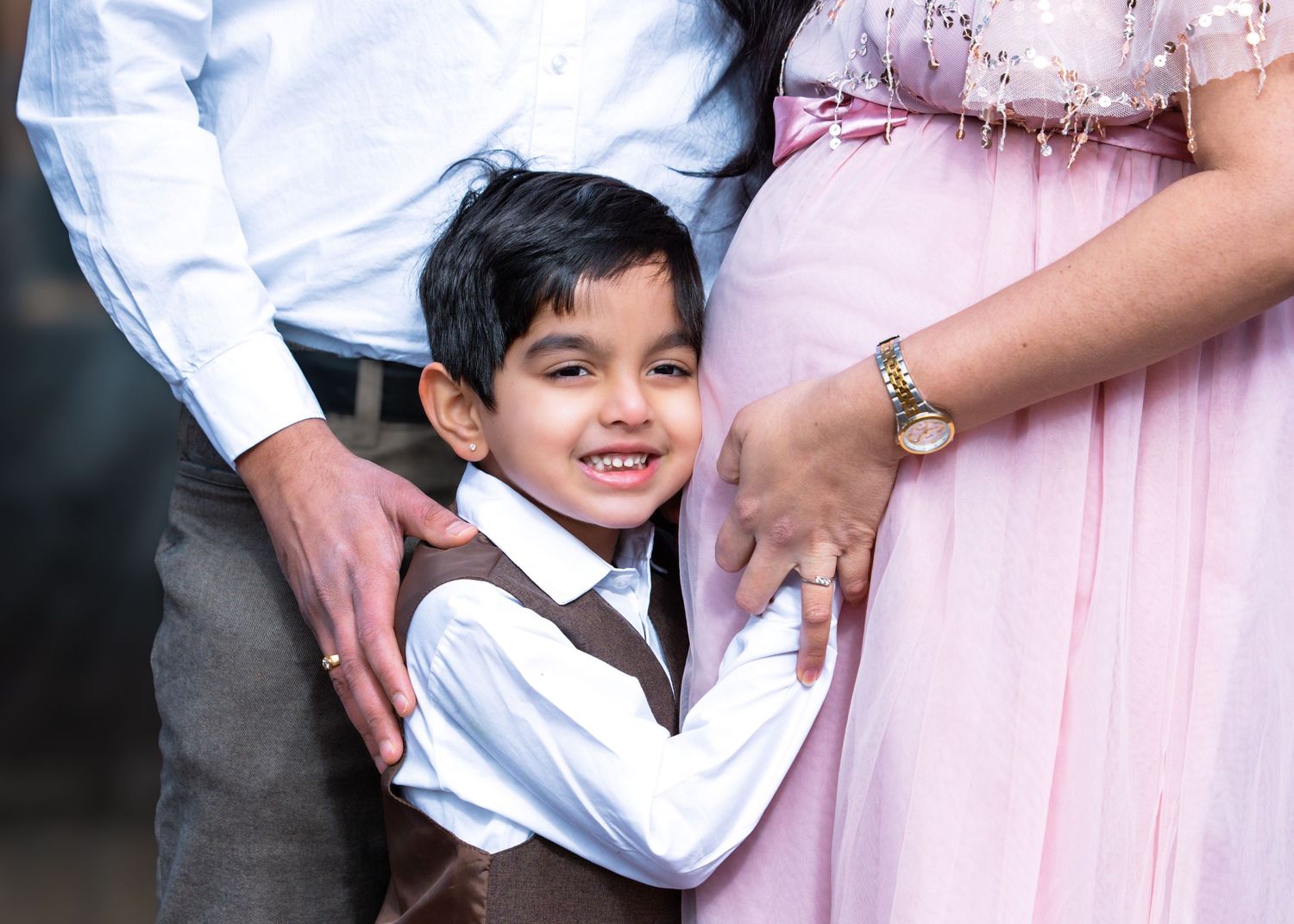 Young boy kissing pregnant mothers belly while standing between parents during an outdoor maternity photoshoot.
