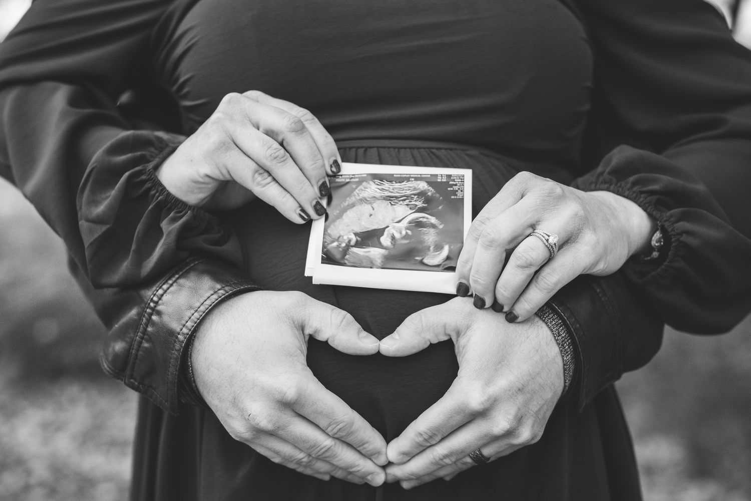 Black and white photo of couple holding ultrasound scan over pregnant belly with heart hands.