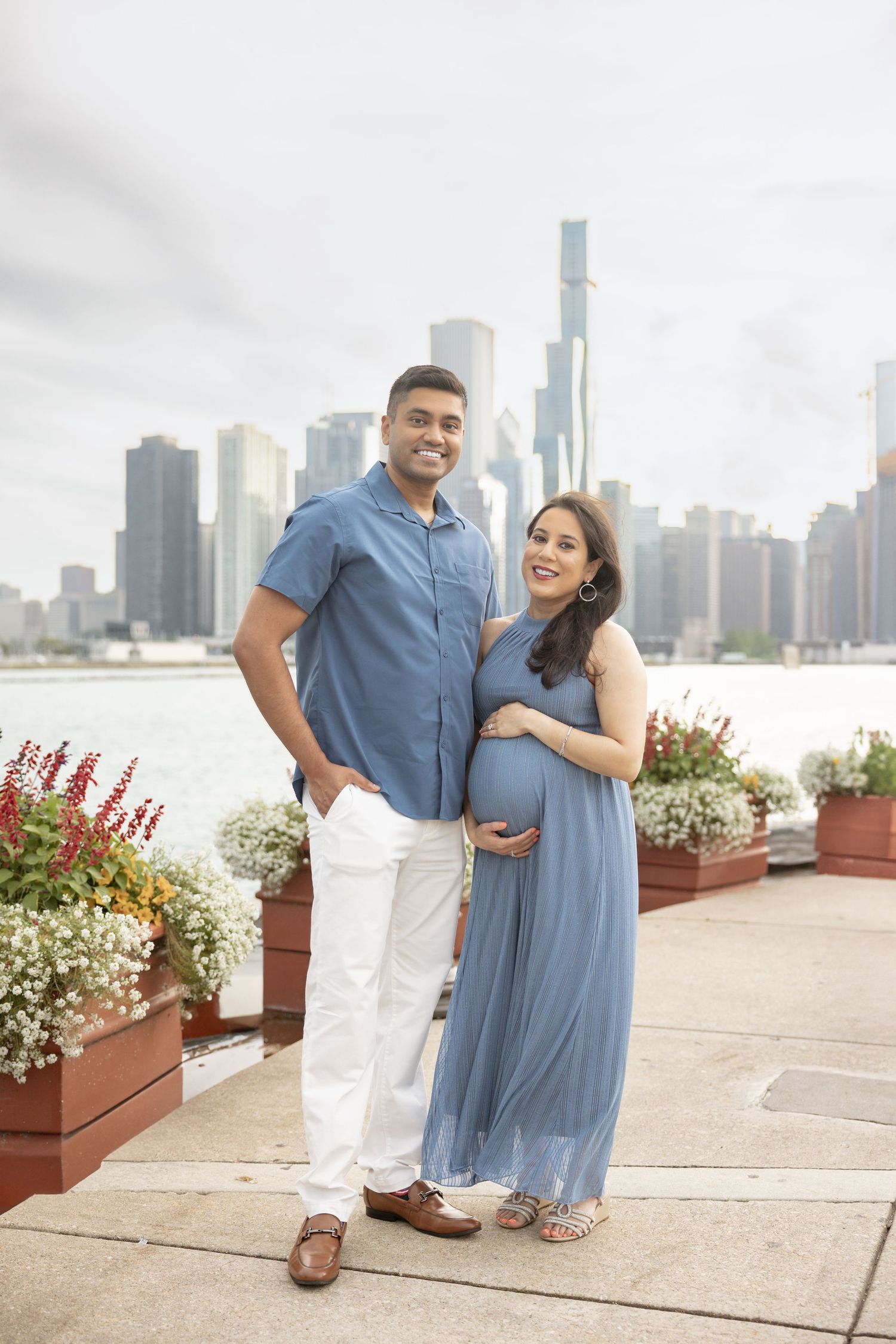 Expecting couple posing by Lake Michigan with Chicago skyline, woman in blue maternity dress cradling baby bump.