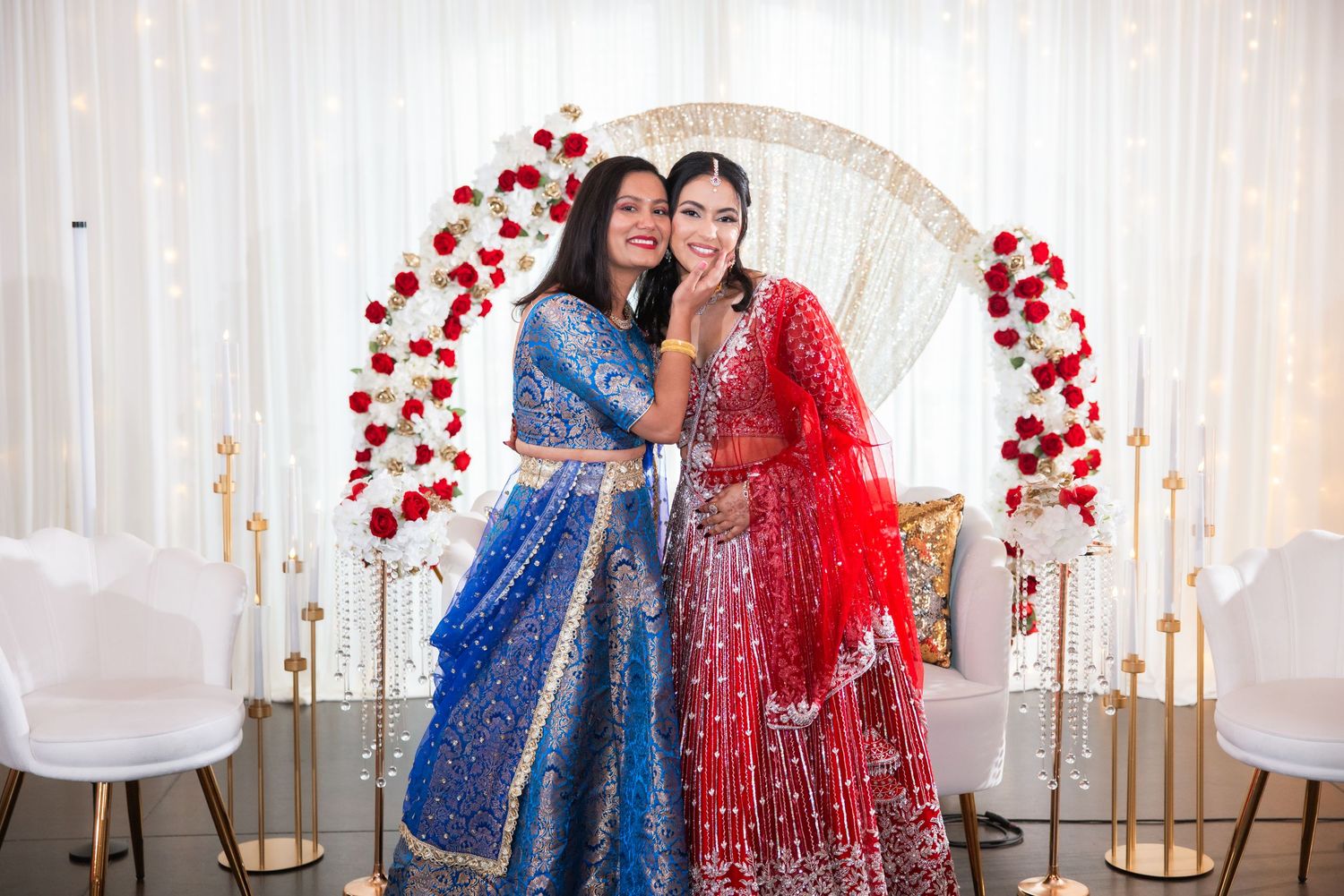 Two women in vibrant blue and red Indian lehenga outfits posing together at a decorated wedding venue.
