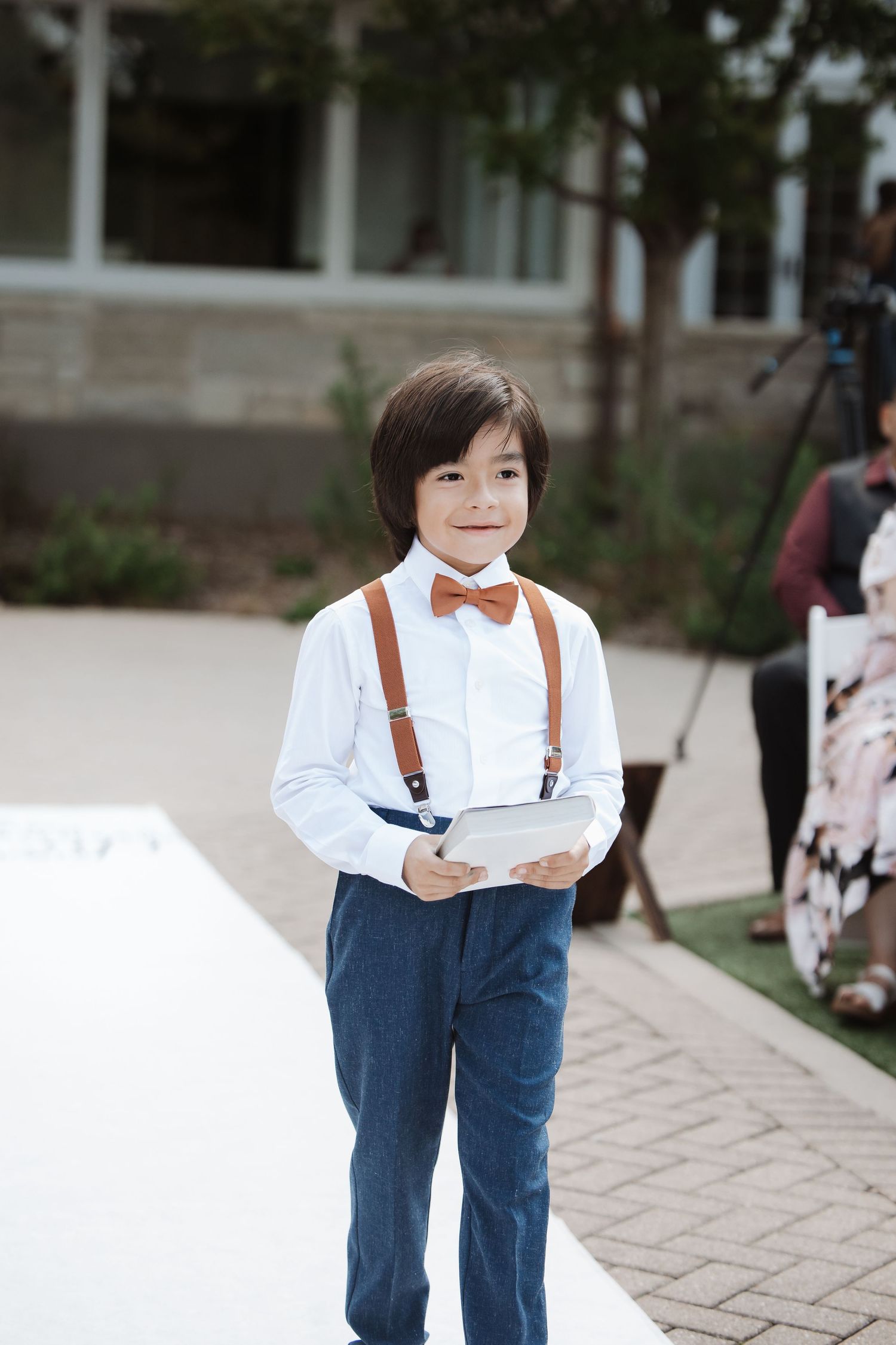 Young ring bearer in white shirt, suspenders, and brown bow tie holds pillow at wedding.
