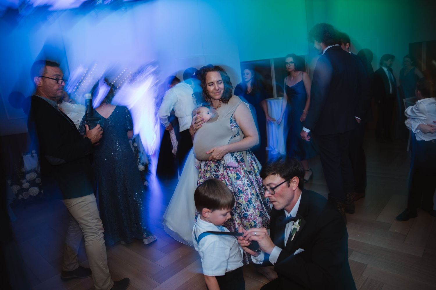 Wedding guests including children dance together under colorful green and blue lights at a lively reception.