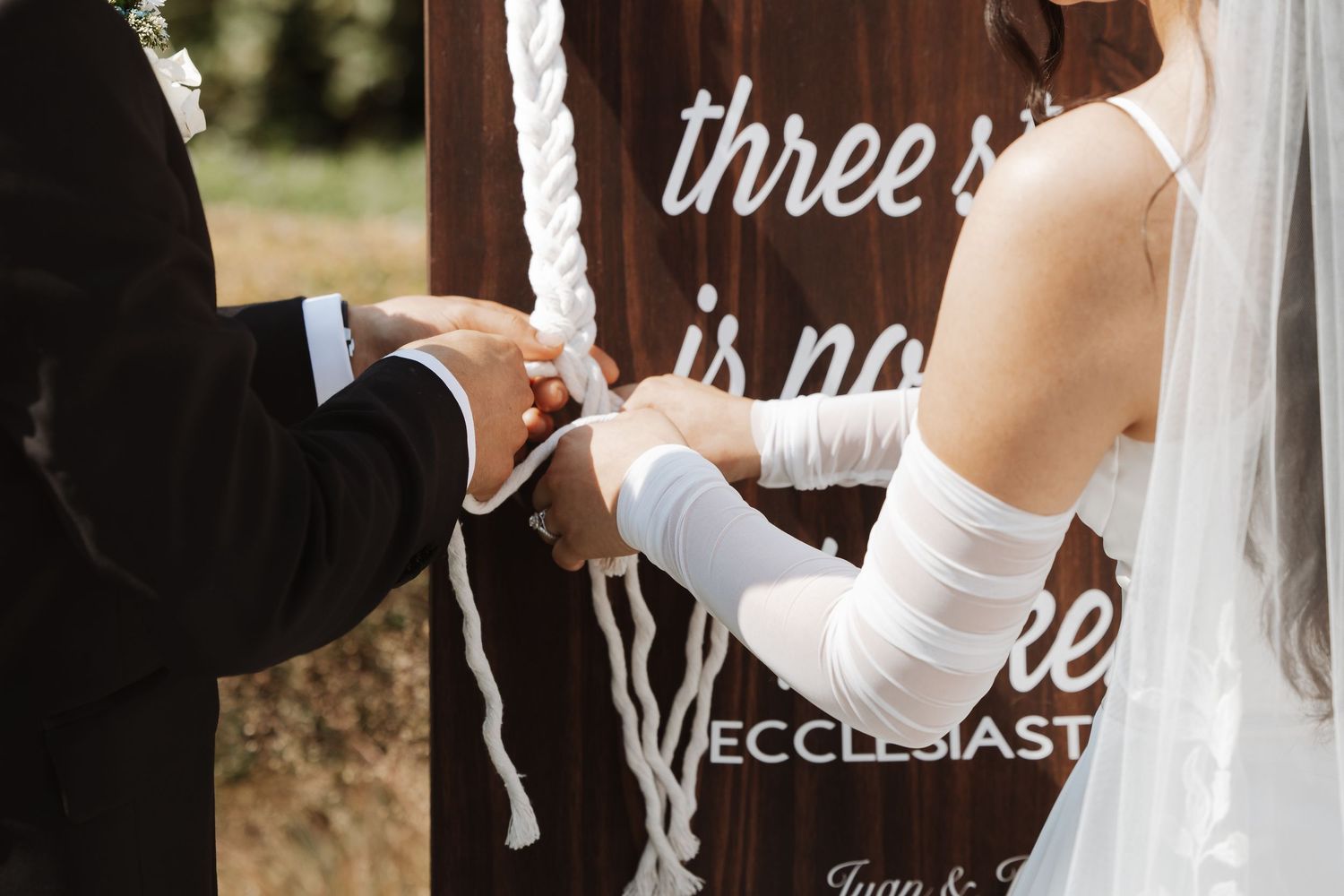 Bride and groom braiding unity cord together during outdoor wedding ceremony at wooden sign.