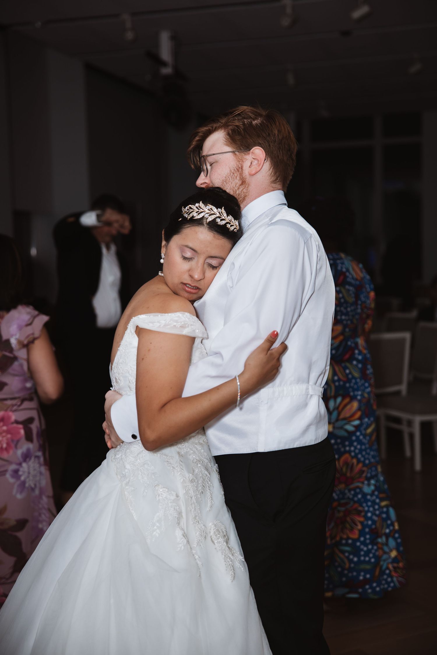 A bride in a white gown and tiara shares a tender first dance embrace with her groom at their wedding reception.