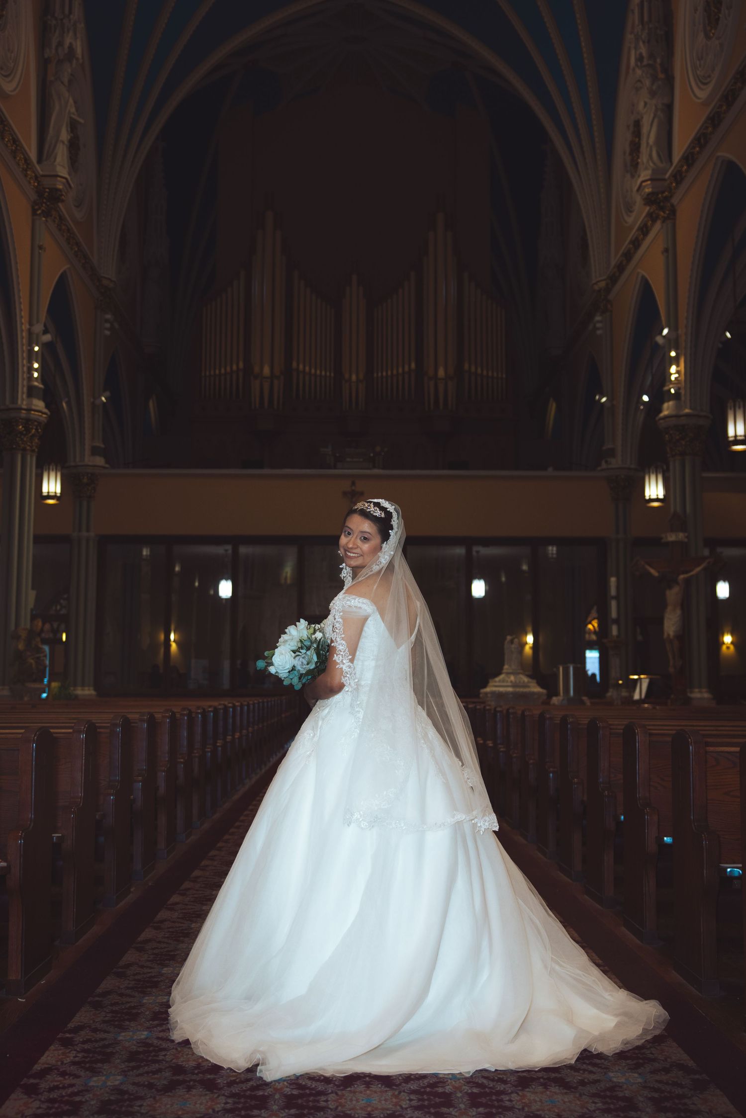 Bride in white ball gown with cathedral veil poses in church aisle with grand pipe organ background.