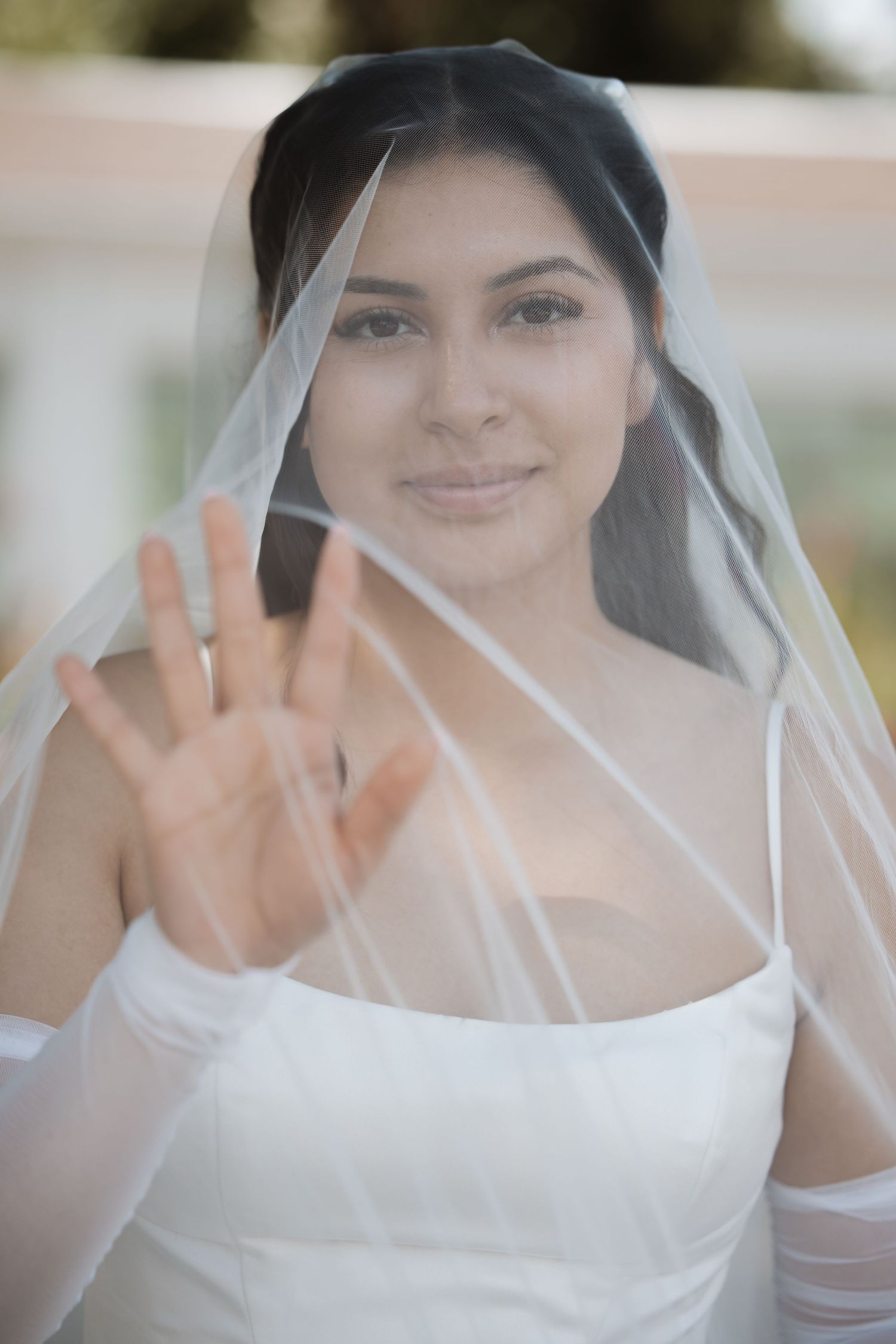 Bride in white wedding dress and veil holds hand up toward camera, smiling softly.