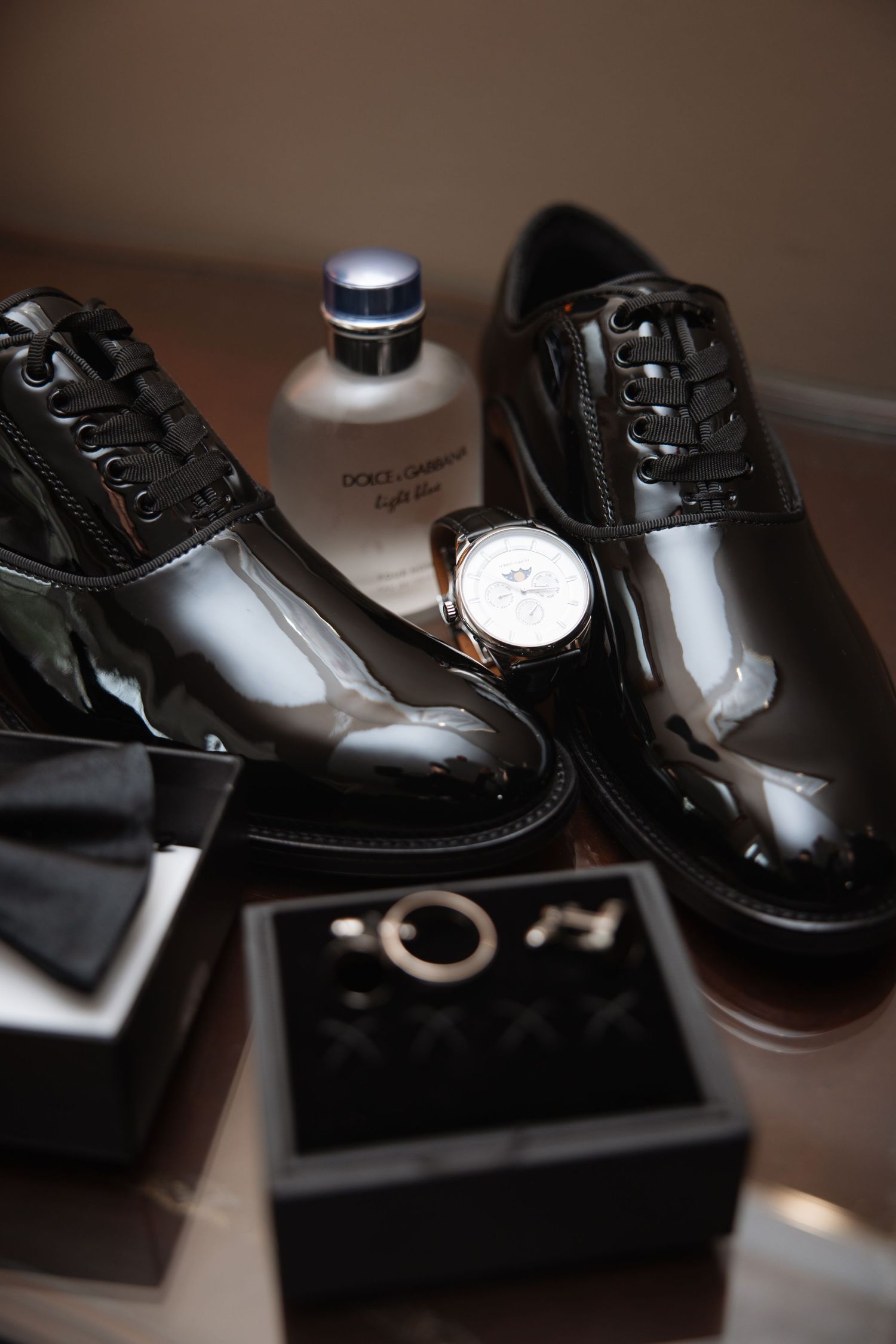 Black patent leather dress shoes with watch, bow tie, cufflinks, and Dolce Gabbana cologne for wedding day groom prep.