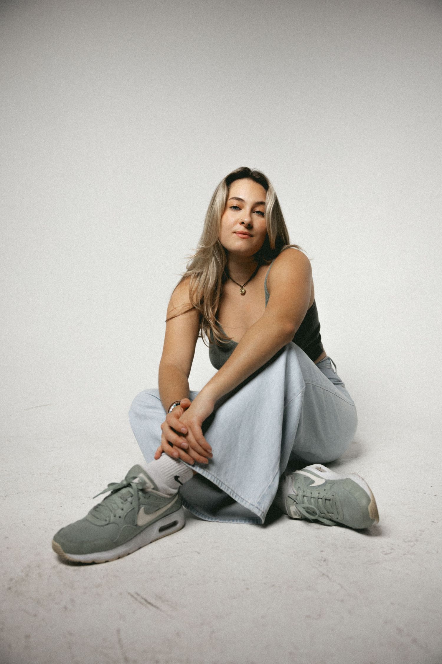 Blonde woman in olive tank top and light jeans sits cross-legged on floor wearing gray Nike sneakers.