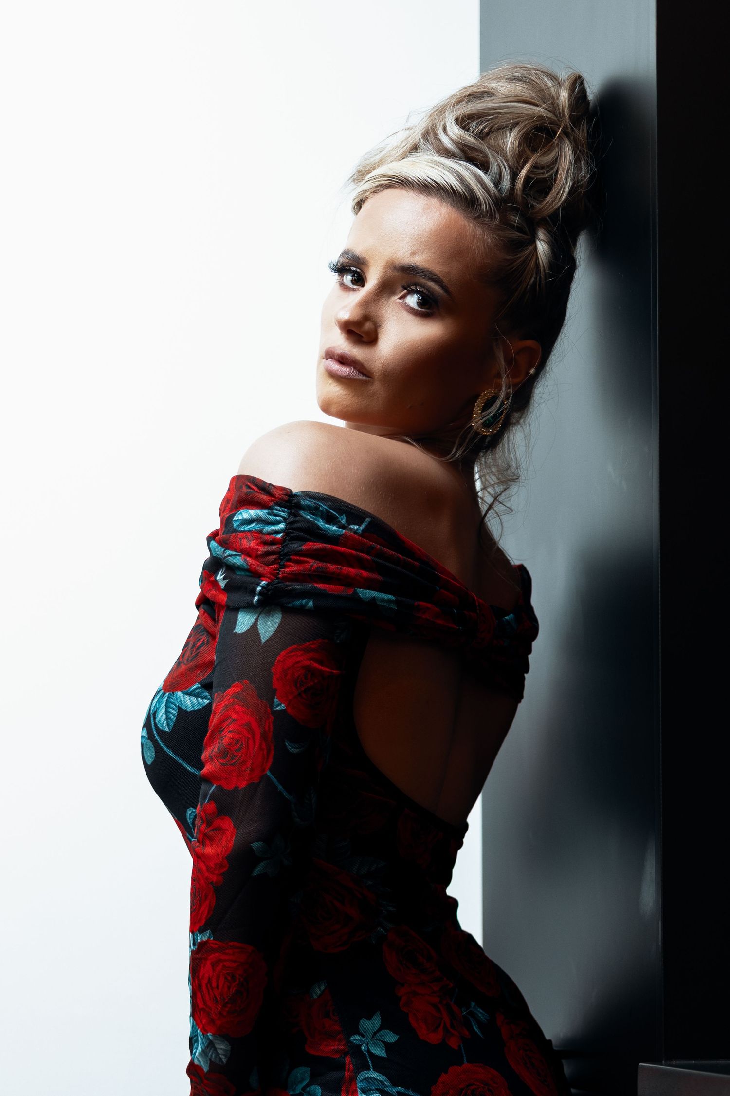 Blonde woman with updo in off-shoulder red floral dress leaning against dark wall.