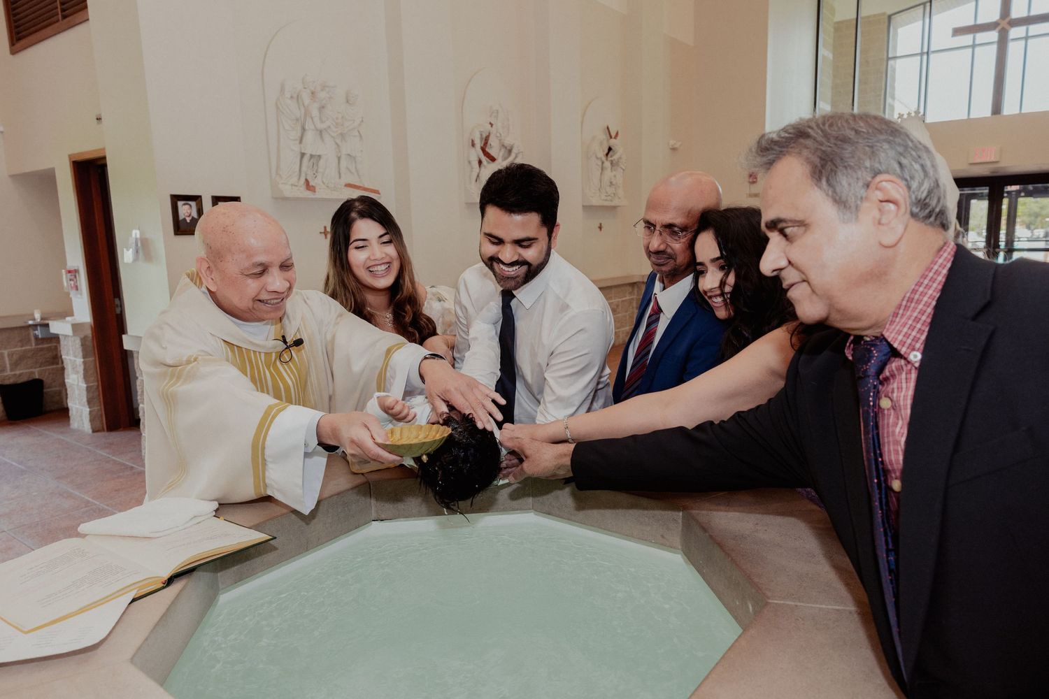 Family gathers at baptismal font with priest during Catholic christening ceremony.