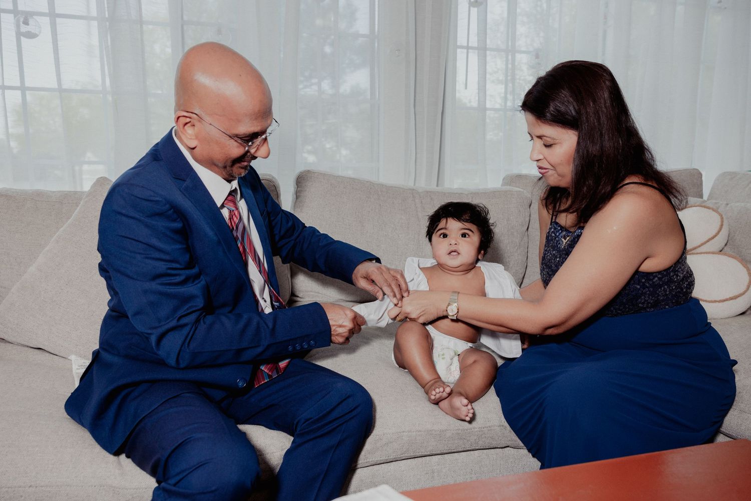 Grandparents sitting on sofa interacting with baby during christening day preparations at home.