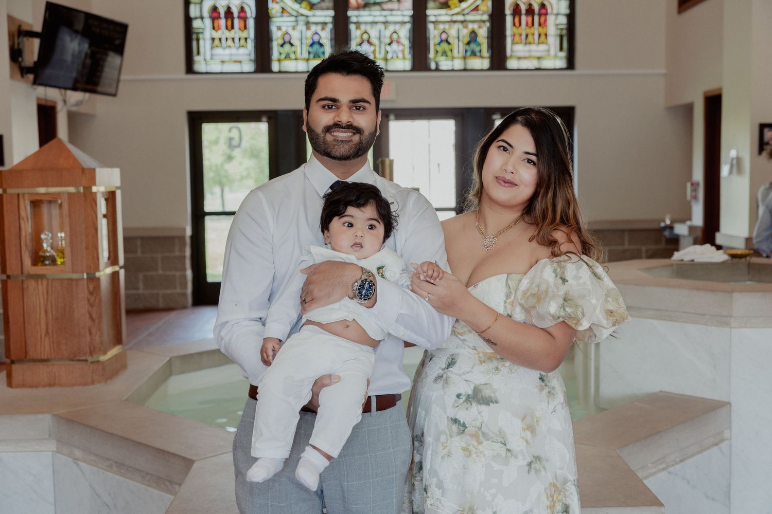 Family of three posing by baptismal font inside church during baby christening ceremony.