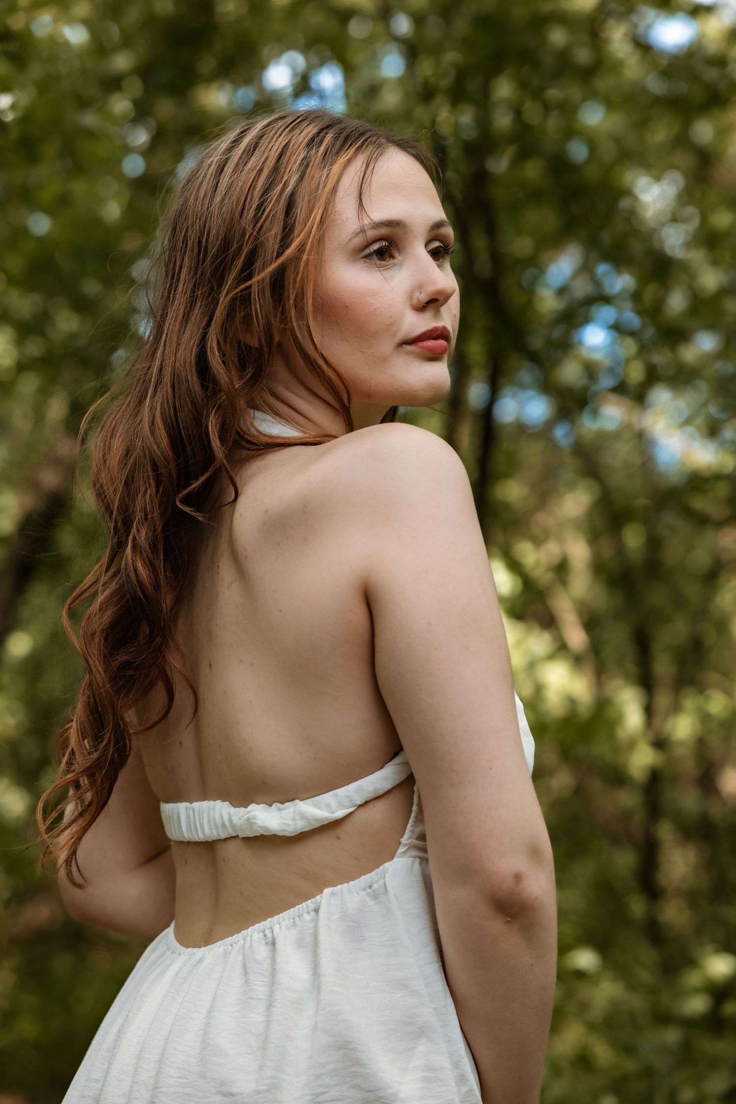 Woman with long auburn hair in backless white dress looks over shoulder in forest.