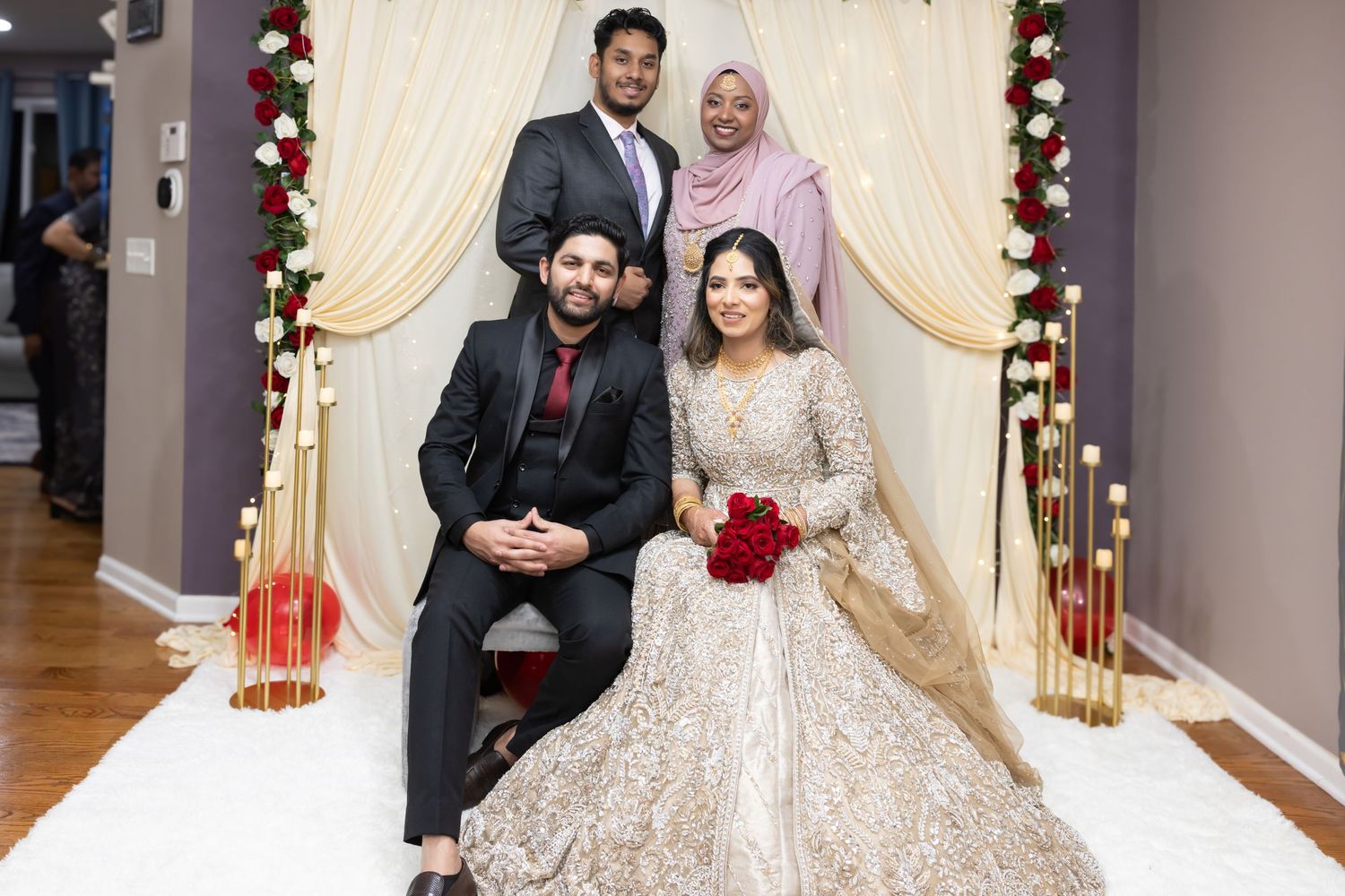 South Asian bride and groom posing with two guests at floral decorated wedding mandap.