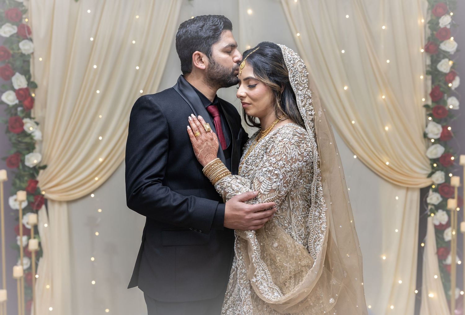 A groom in a black suit kisses his bride wearing a traditional embroidered wedding dress at a decorated altar.