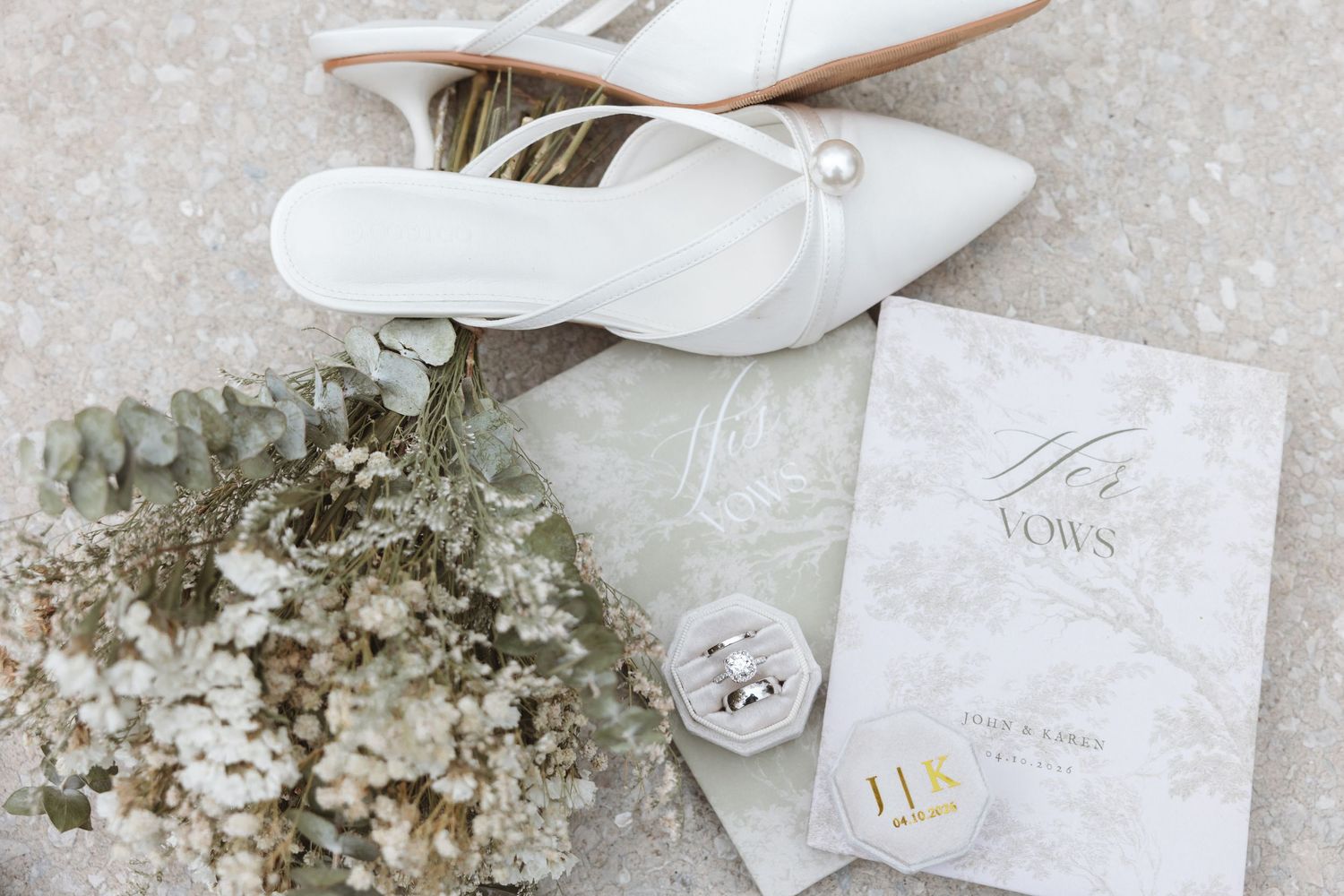 White wedding heels, floral bouquet, rings, and vow booklets arranged elegantly on a neutral surface.