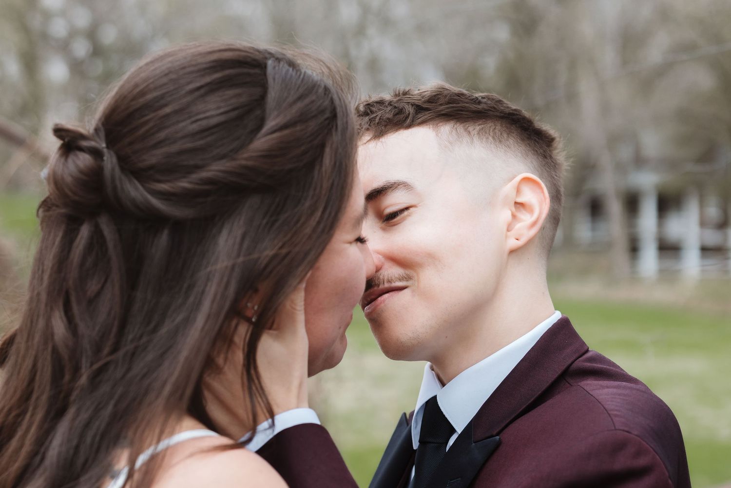 Two brides sharing a romantic kiss outdoors during their wedding ceremony, one in a burgundy suit and one in a white dress.