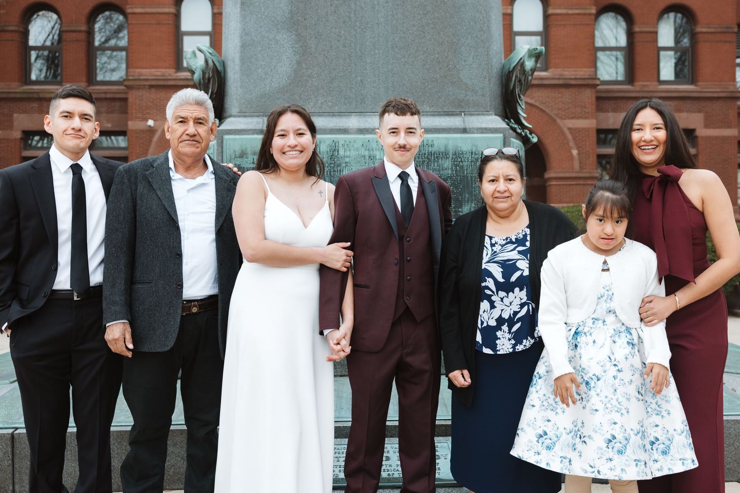 Family group photo of seven people posing outdoors near a statue, dressed in formal attire for a special occasion.