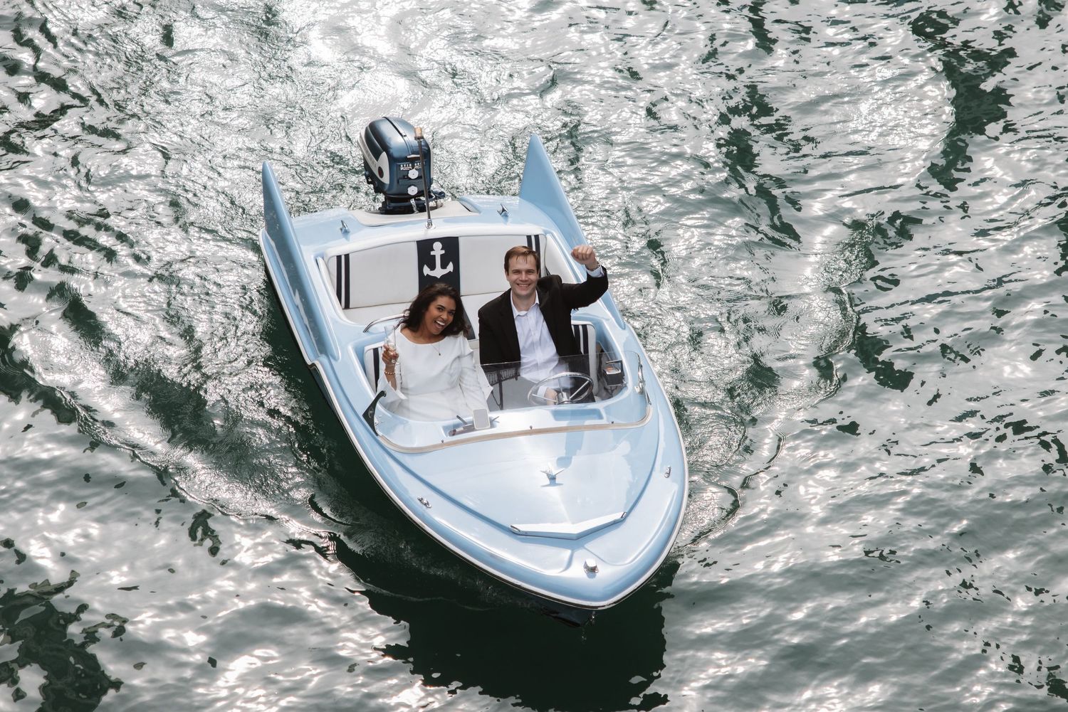 Aerial view of a couple waving from a light blue speedboat surrounded by sparkling water on the Chicago River.