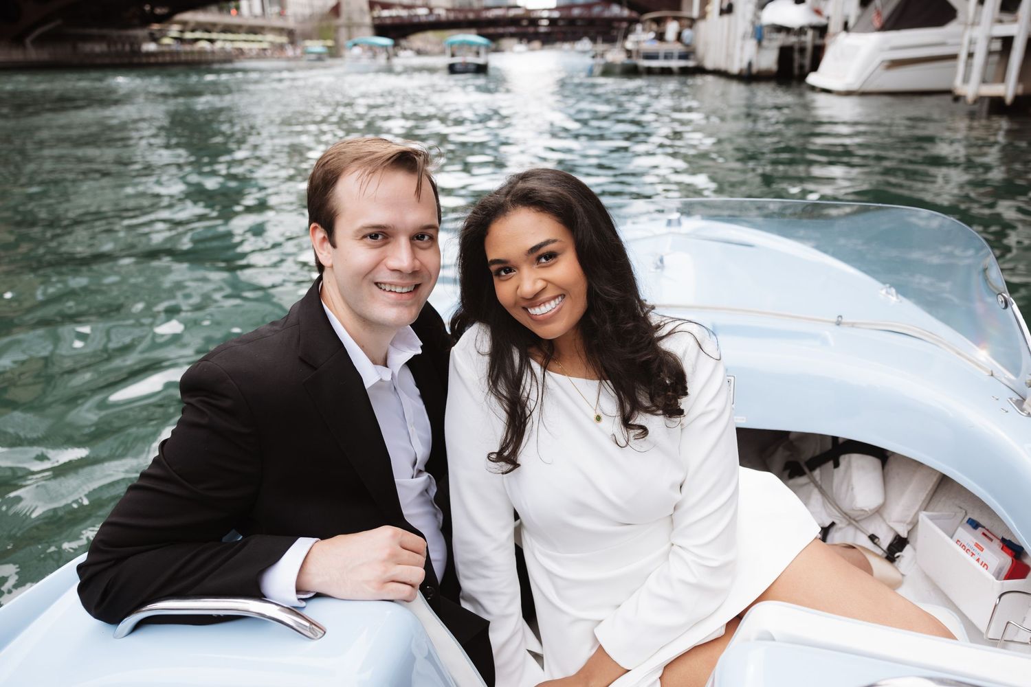 Smiling couple sitting together on a white boat in a marina, man in suit and woman in white outfit.