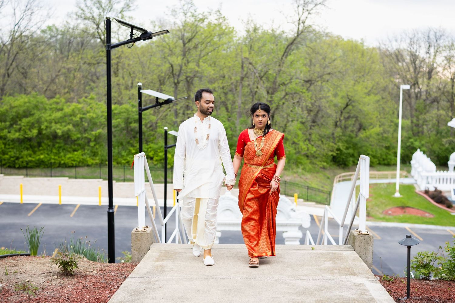 Indian couple in traditional wedding attire holding hands walking on a bridge pathway.