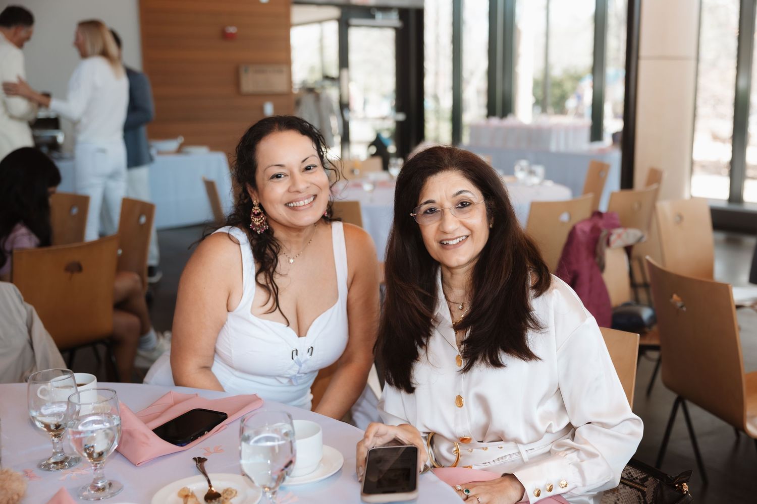 Two smiling women in white attire seated together at an elegant indoor baby shower luncheon event.