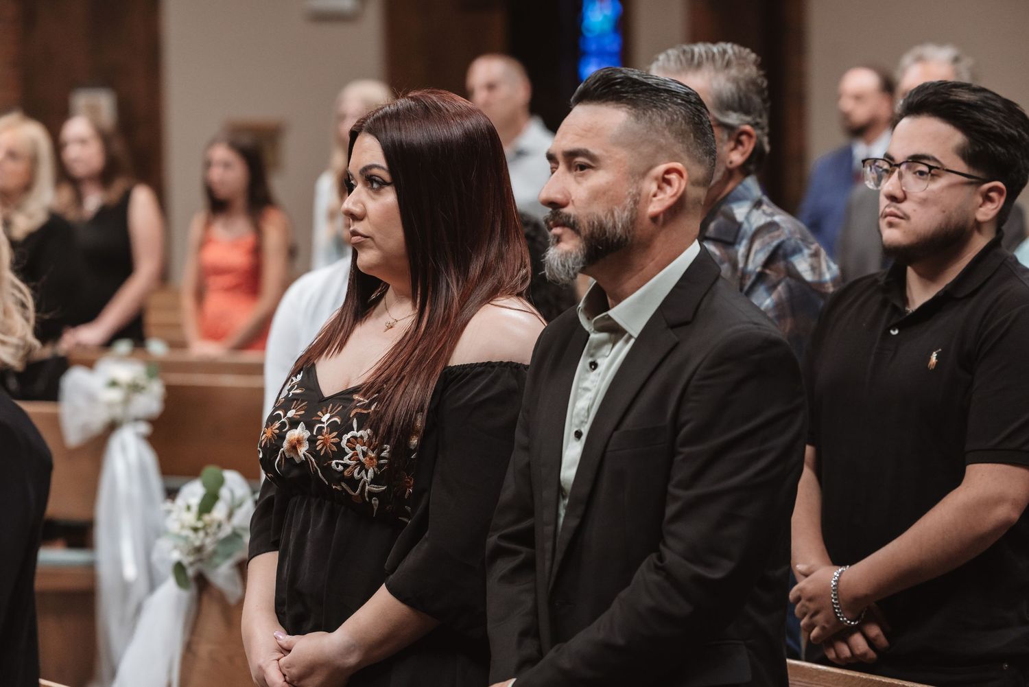 Guests standing in church pews during a wedding ceremony, dressed in formal attire inside an elegant chapel.