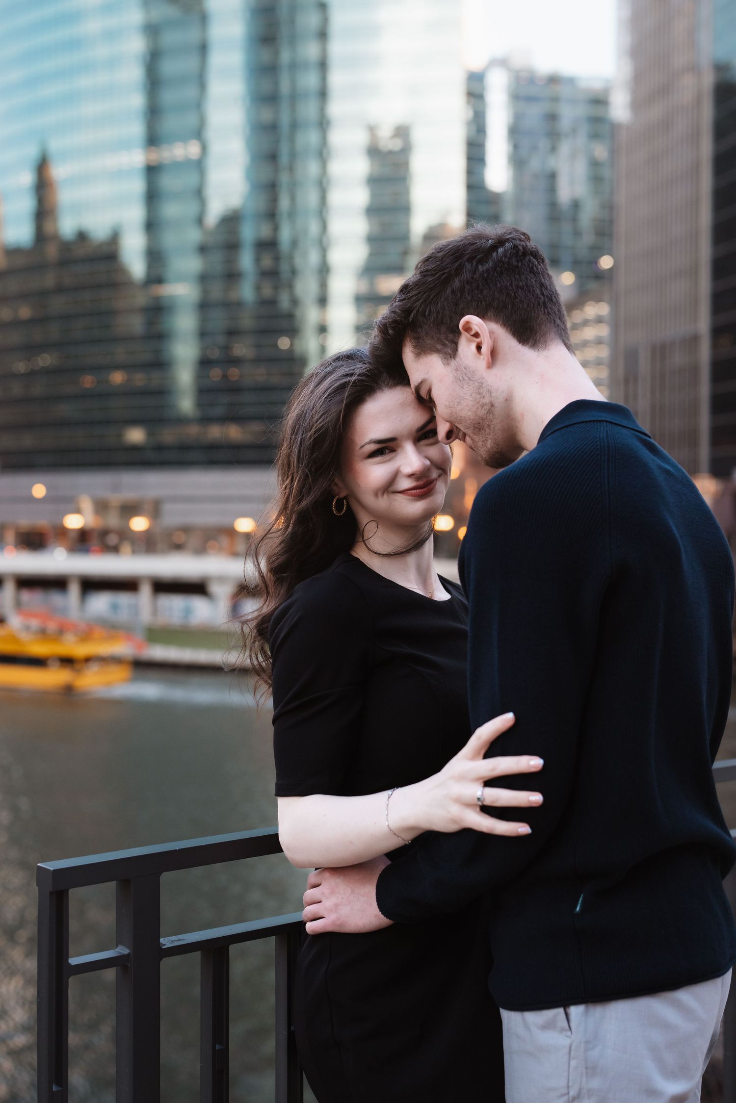 A couple shares a tender embrace on a bridge with Chicago river and city buildings in background.