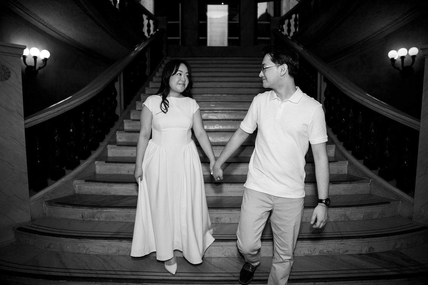 Black and white photo of an engaged couple holding hands and walking down a grand staircase together.