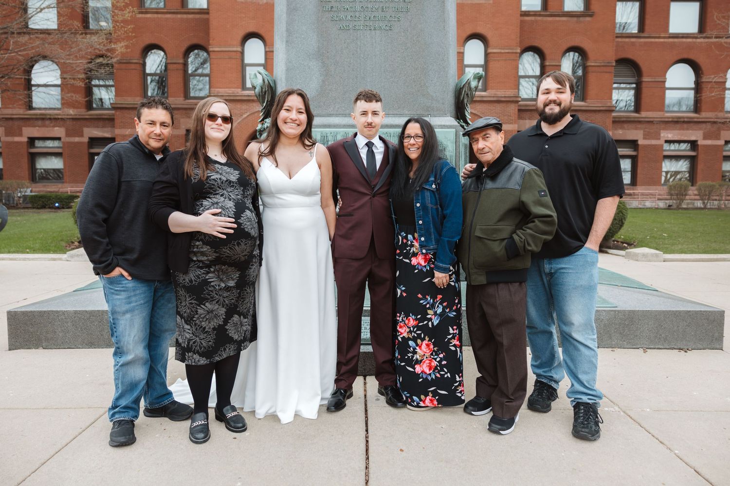 Wedding party of seven poses outdoors near a monument, with bride in white gown and groom in burgundy suit.