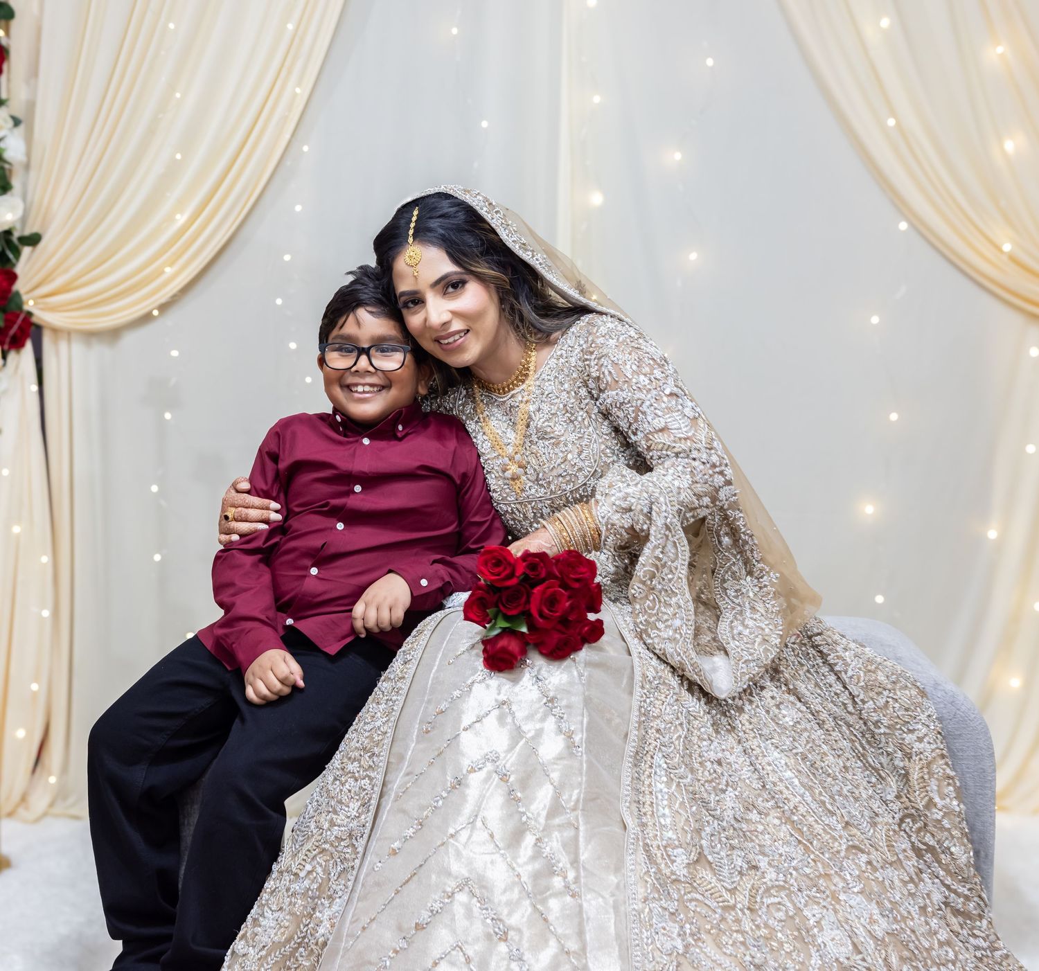 Bride in gold embroidered lehenga holds red roses and poses with young boy in maroon shirt at wedding.